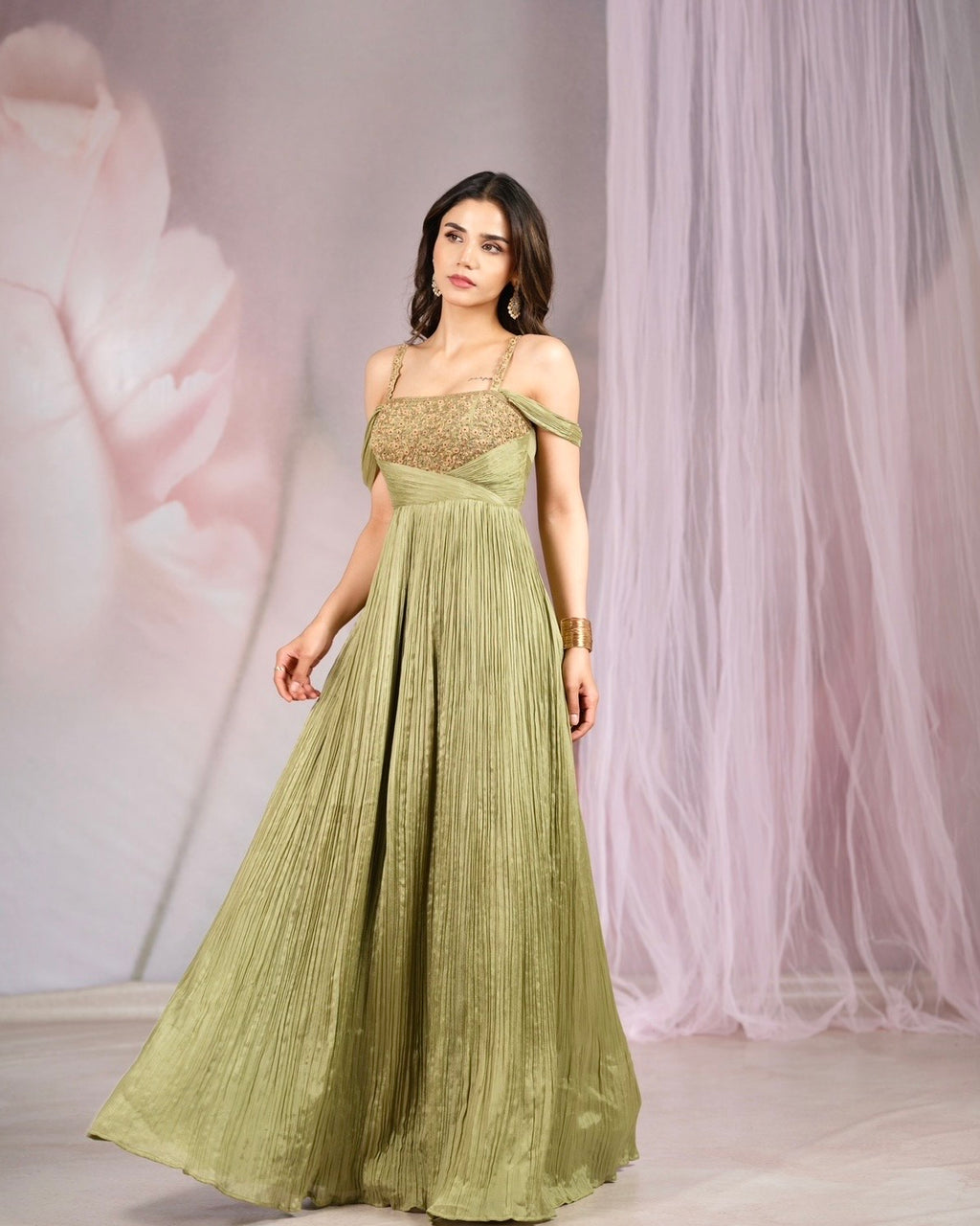 A woman models a floor-length olive green gown with a pleated skirt, a gold embroidered bodice, and off-the-shoulder straps, set against a soft pink and white draped backdrop.