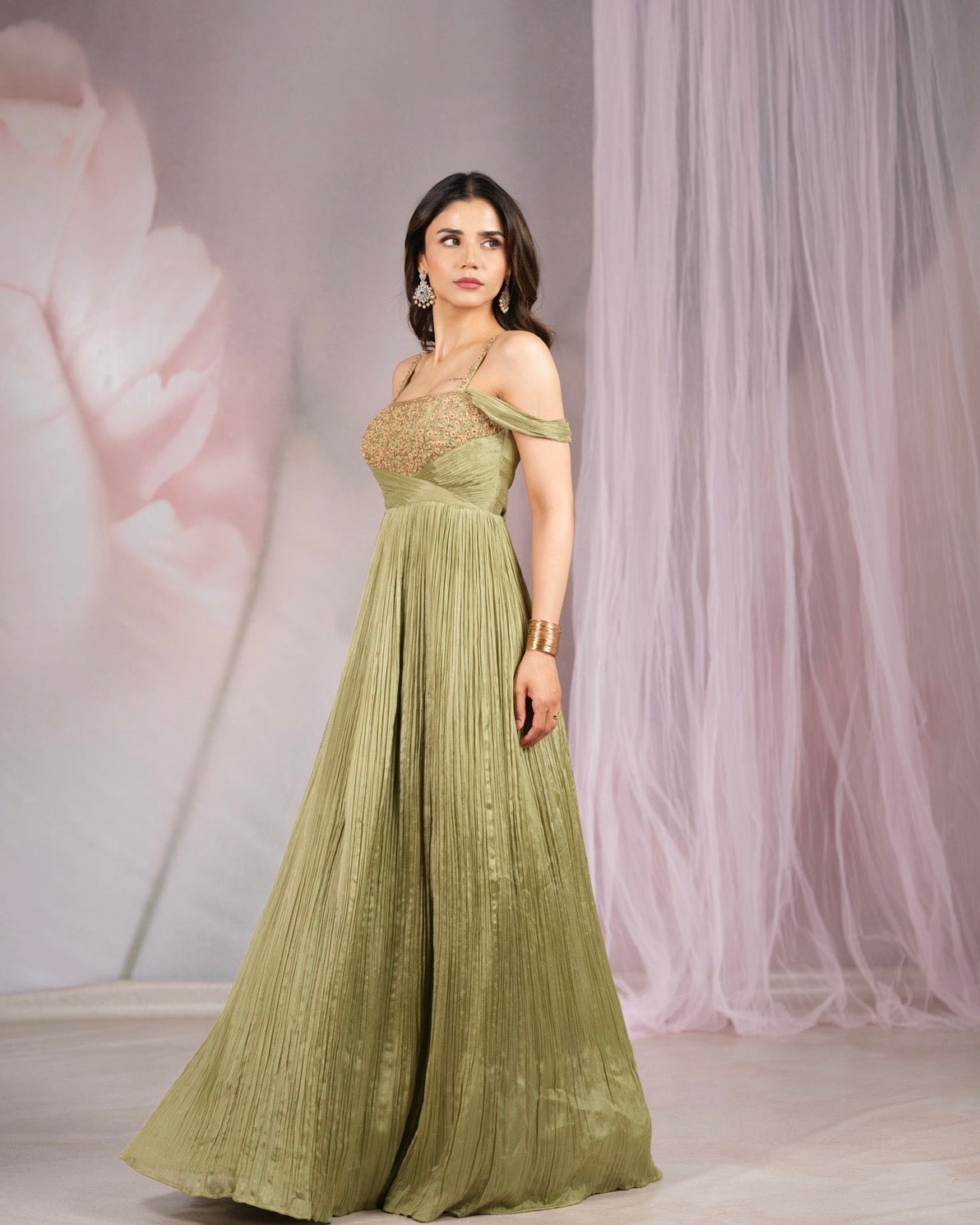 A woman modeling a floor-length olive green gown with off-the-shoulder straps, a lace bodice, and a pleated skirt, posed against a soft pink and white backdrop.
