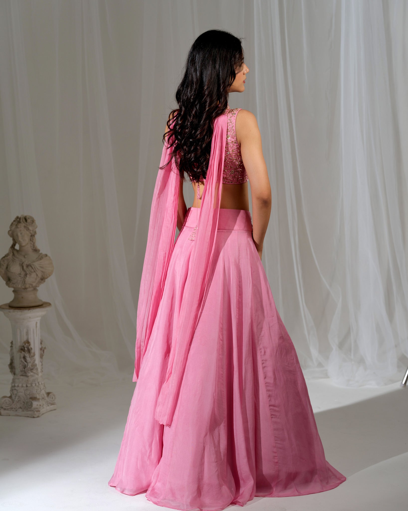 Woman in a pink saree standing against a white curtain backdrop