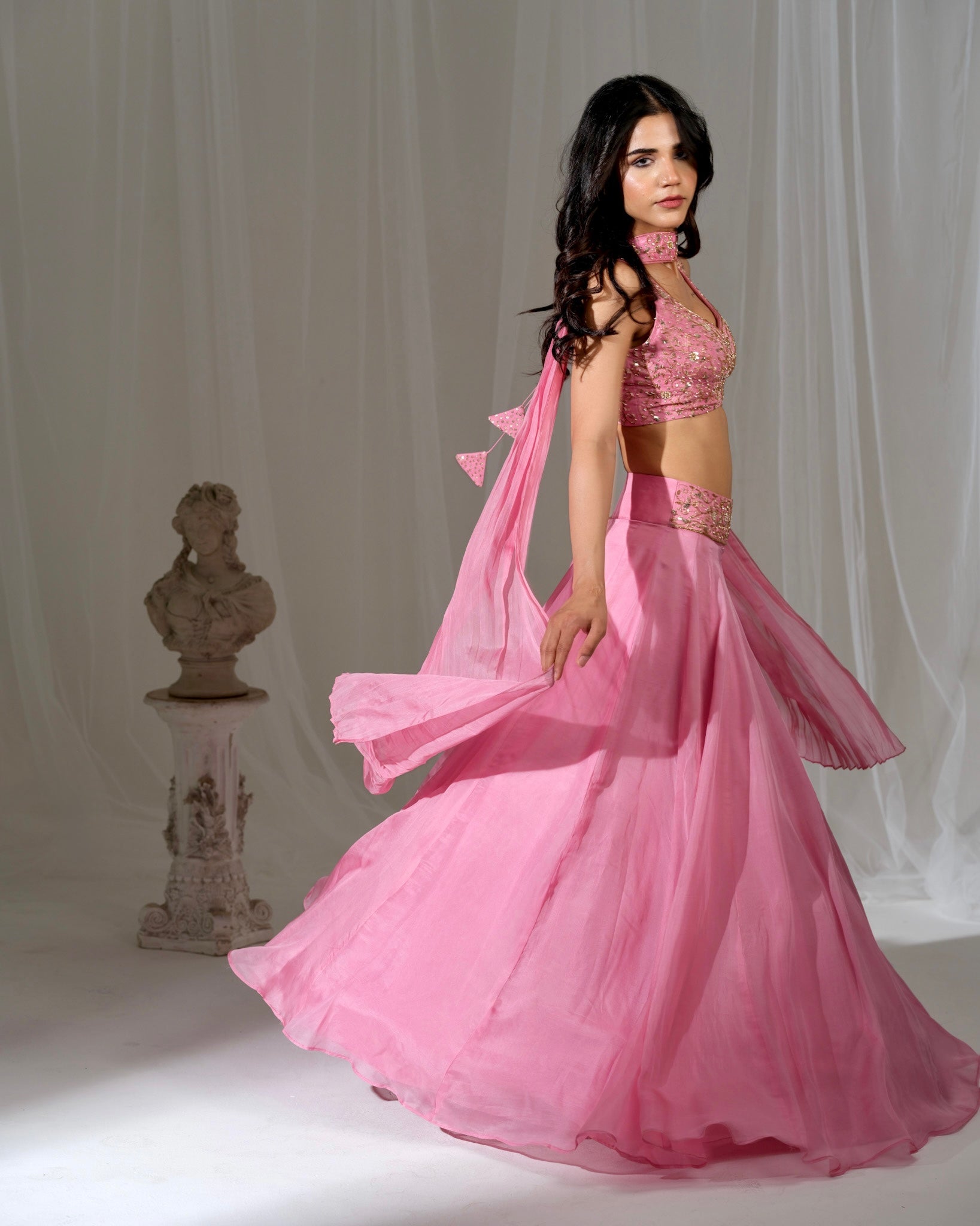 Woman in a pink dress standing against a white curtain with a bust on a pedestal.