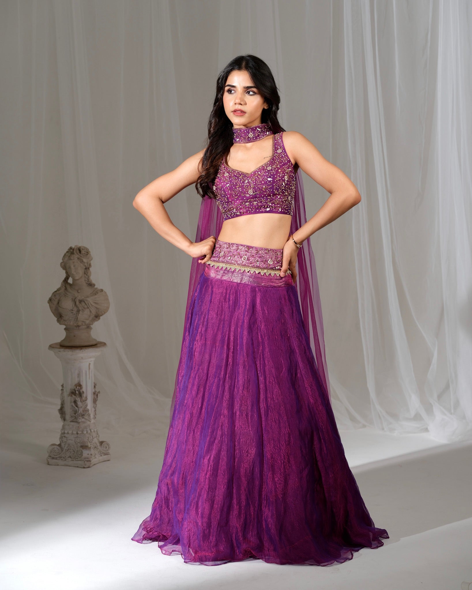 Woman wearing a purple traditional outfit with a sheer dupatta against a white curtain background