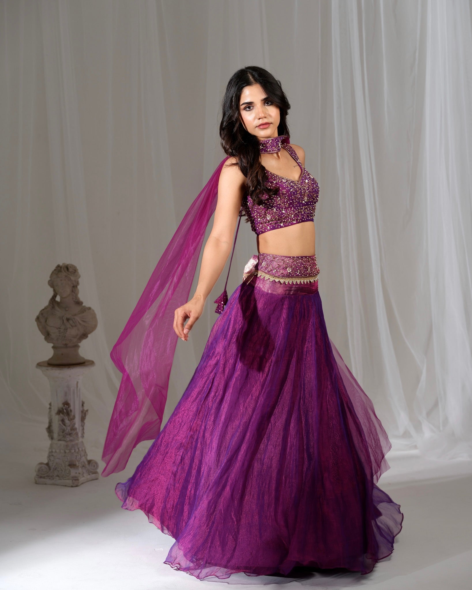 Woman wearing a purple traditional outfit with a sheer dupatta against a gray background