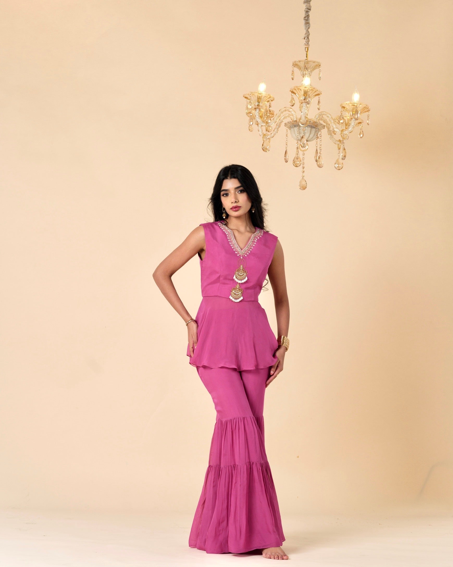 Woman in a pink traditional outfit standing in front of a beige wall with a chandelier.