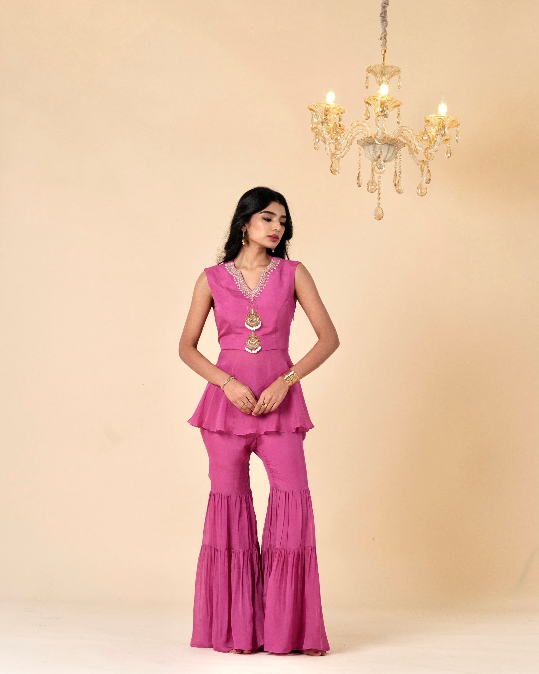 Woman wearing a pink outfit with a beige background and chandelier.