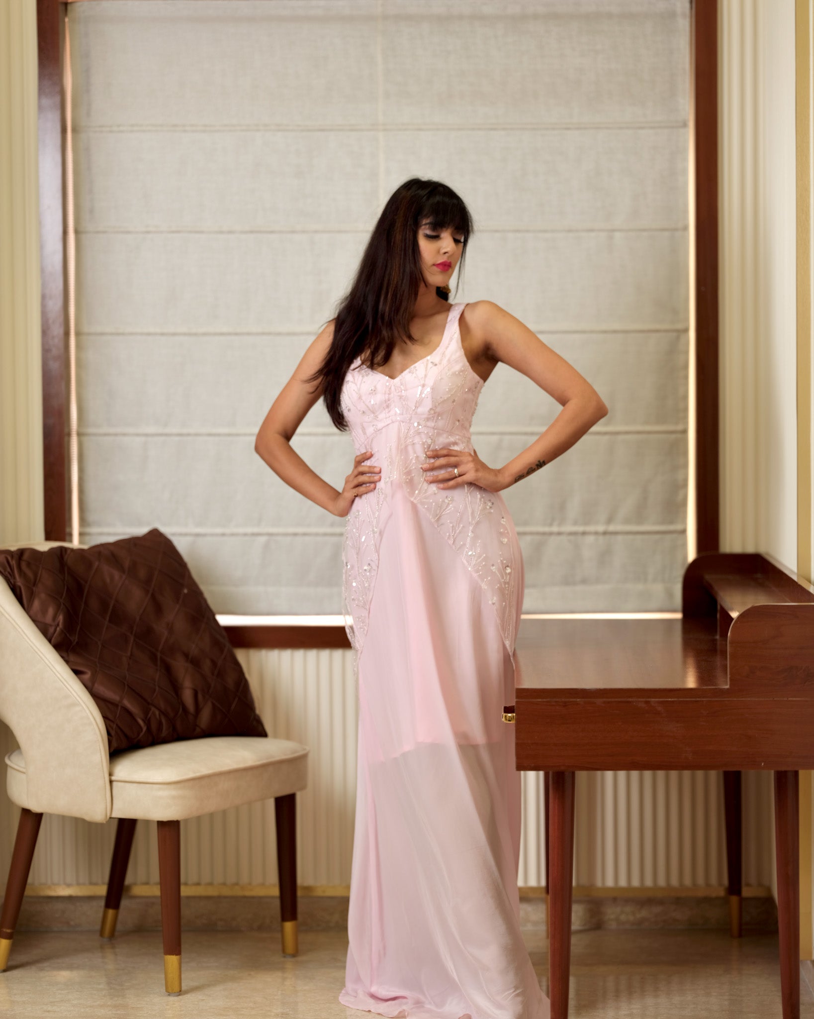 Woman in a pink evening gown standing in a room with a chair and table.