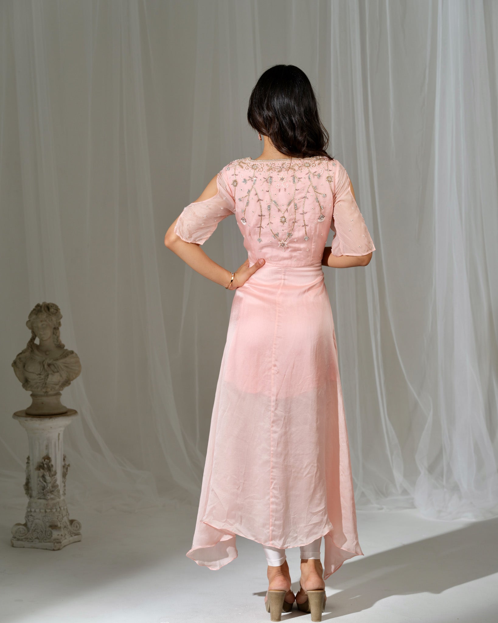Woman wearing a pink dress with lace details, standing against a white curtain.