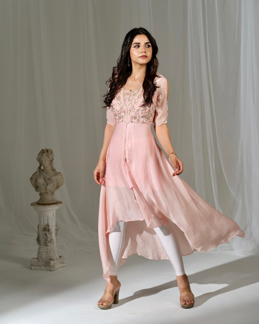 Woman wearing a pink dress with lace top and flowing skirt against a white curtain background.