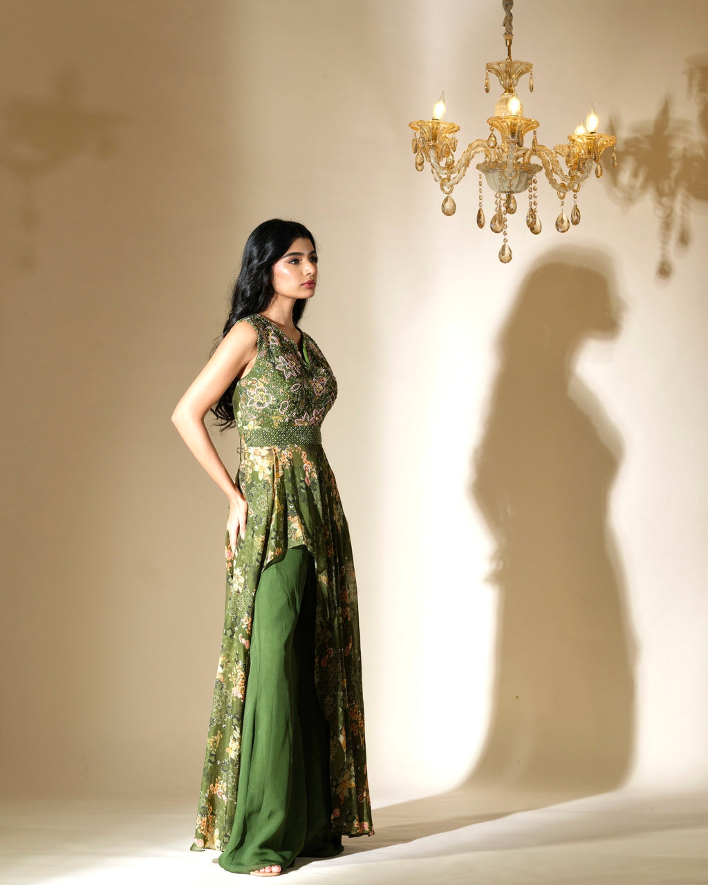 Woman in a green dress standing in front of a chandelier casting a shadow on the wall.