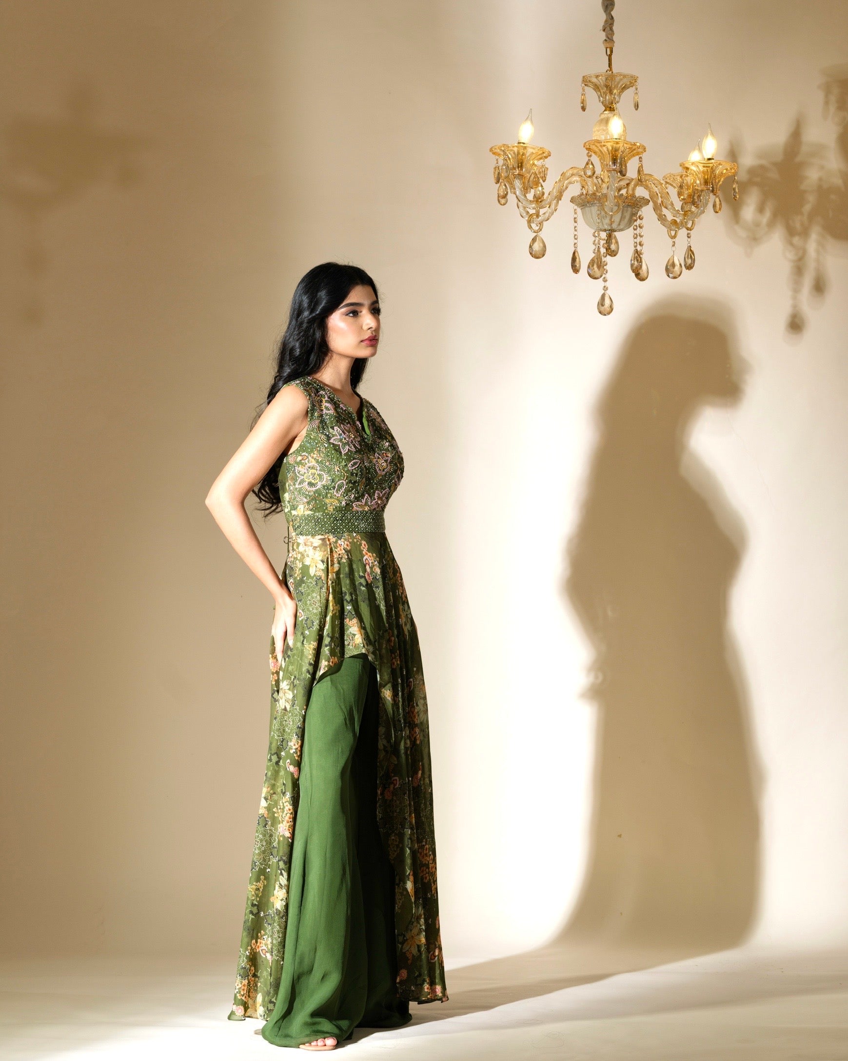 Woman in a green dress standing in front of a chandelier casting a shadow on the wall.