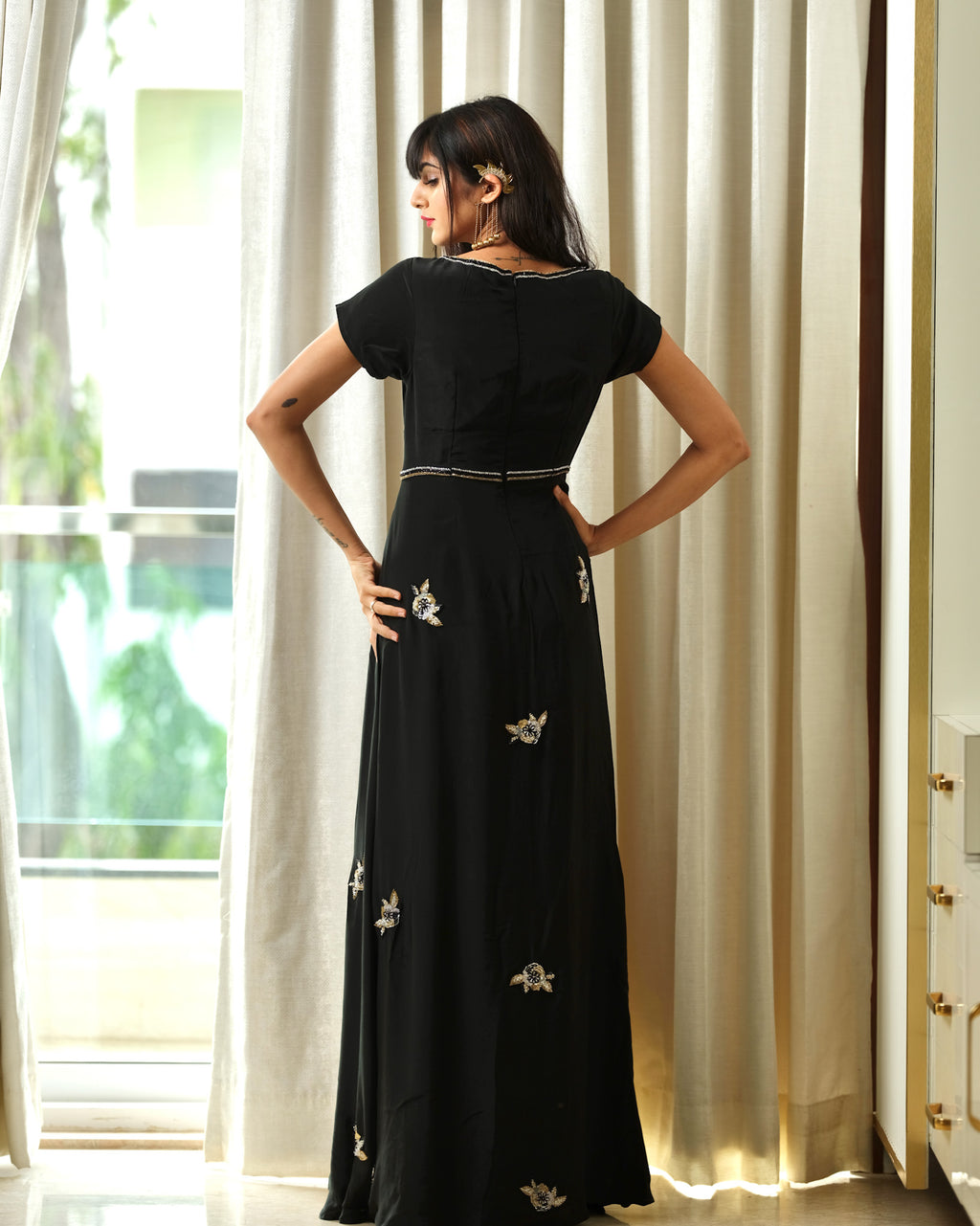 Woman wearing a black dress with floral embellishments standing in a room with curtains.