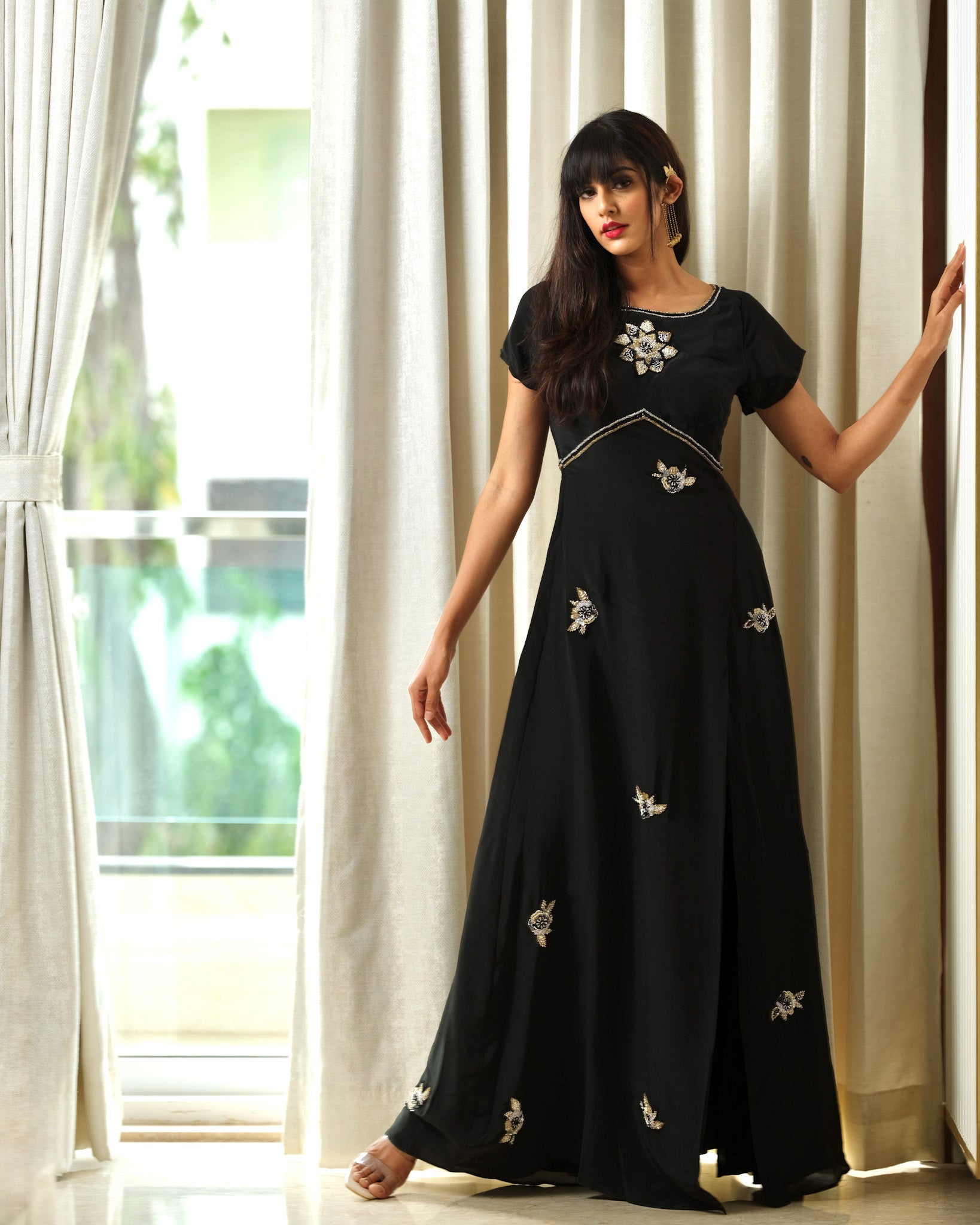 Woman wearing a black dress with floral embroidery in a bright room with white curtains.
