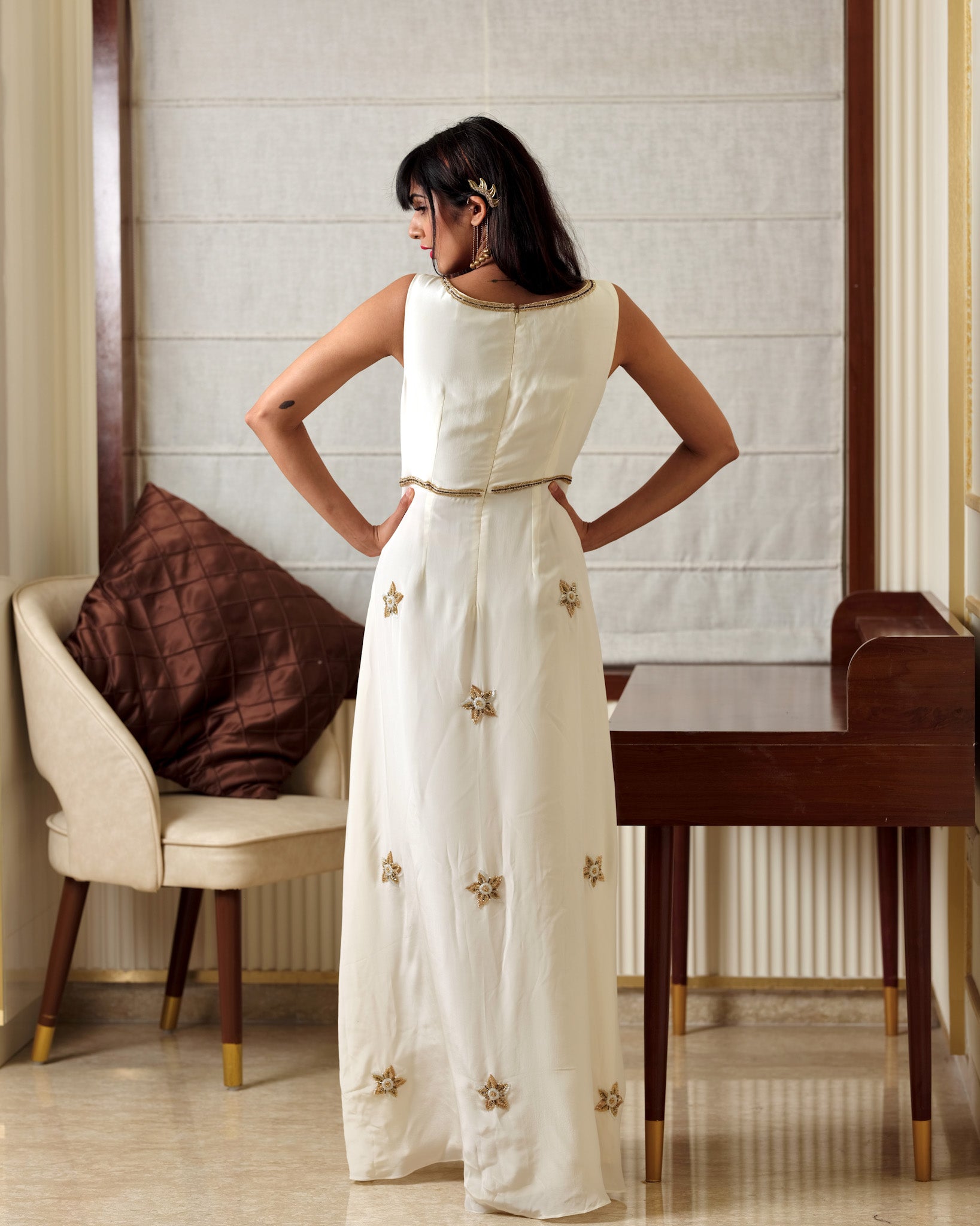 Woman in a white dress with gold embellishments standing in a room with a chair and table.