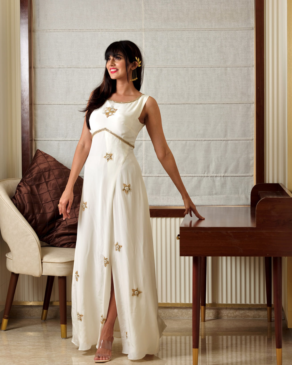 Woman in a white dress with gold accents standing in a room with a chair and table.