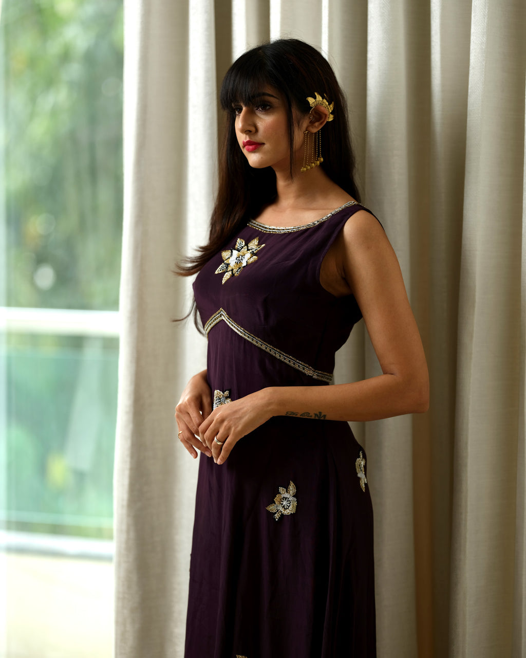 Woman wearing a dark purple dress with floral embellishments in front of a window with white curtains.