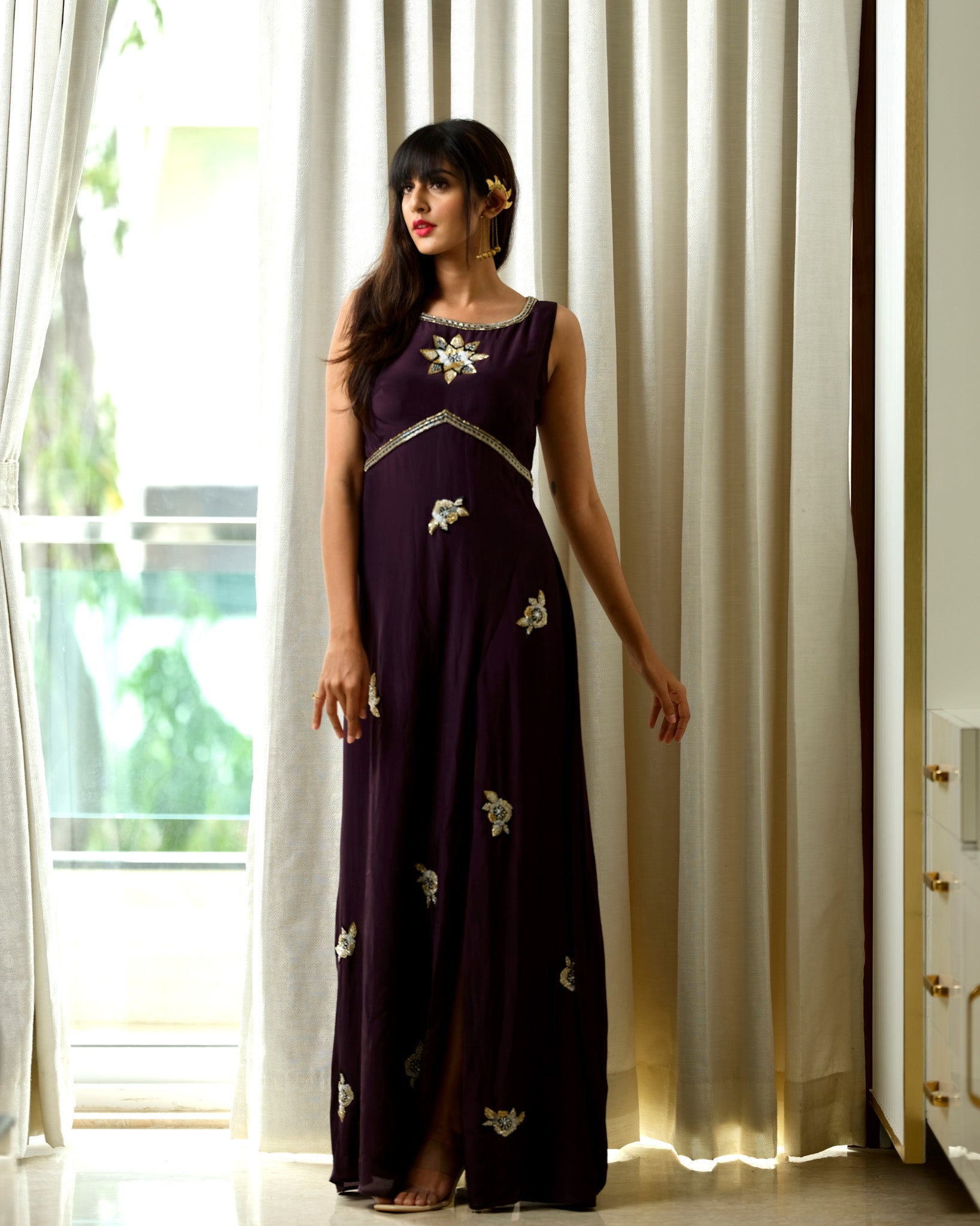 Woman wearing a dark purple dress with floral embellishments in a room with white curtains.