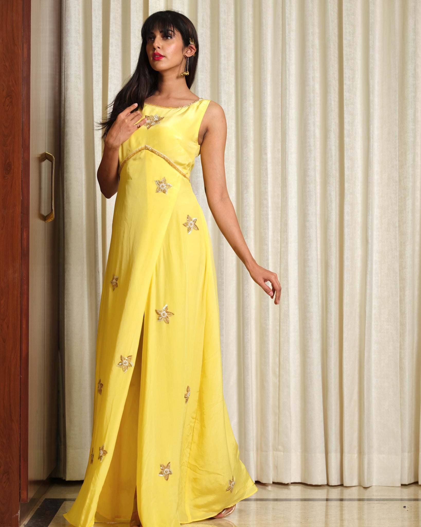 Woman wearing a yellow saree with floral patterns in a room with white curtains.