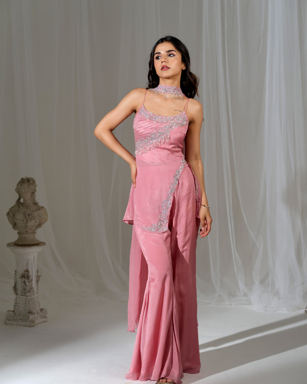 Woman wearing a pink evening gown with silver embellishments against a white curtain background.