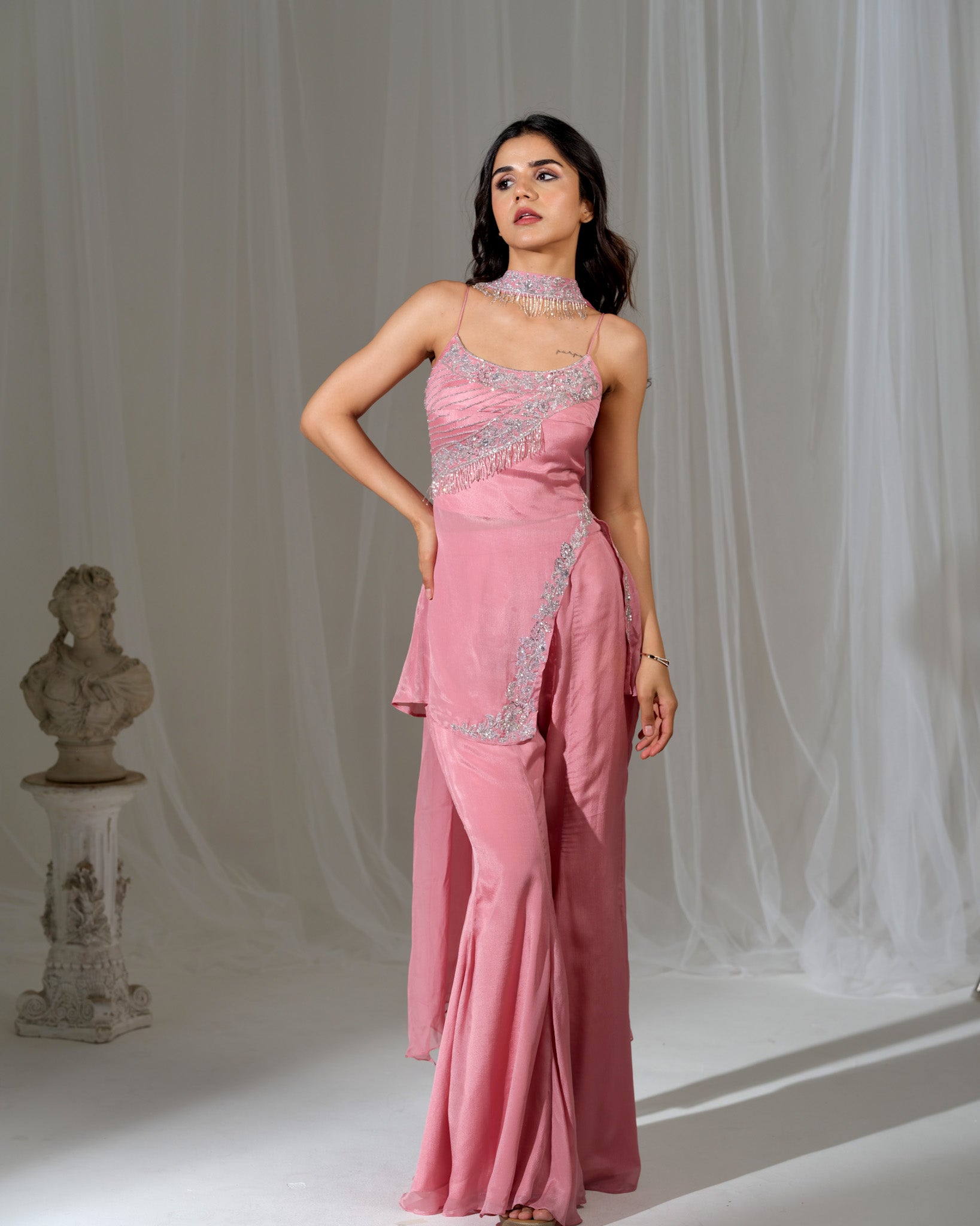 Woman wearing a pink evening gown with silver embellishments against a white curtain background.
