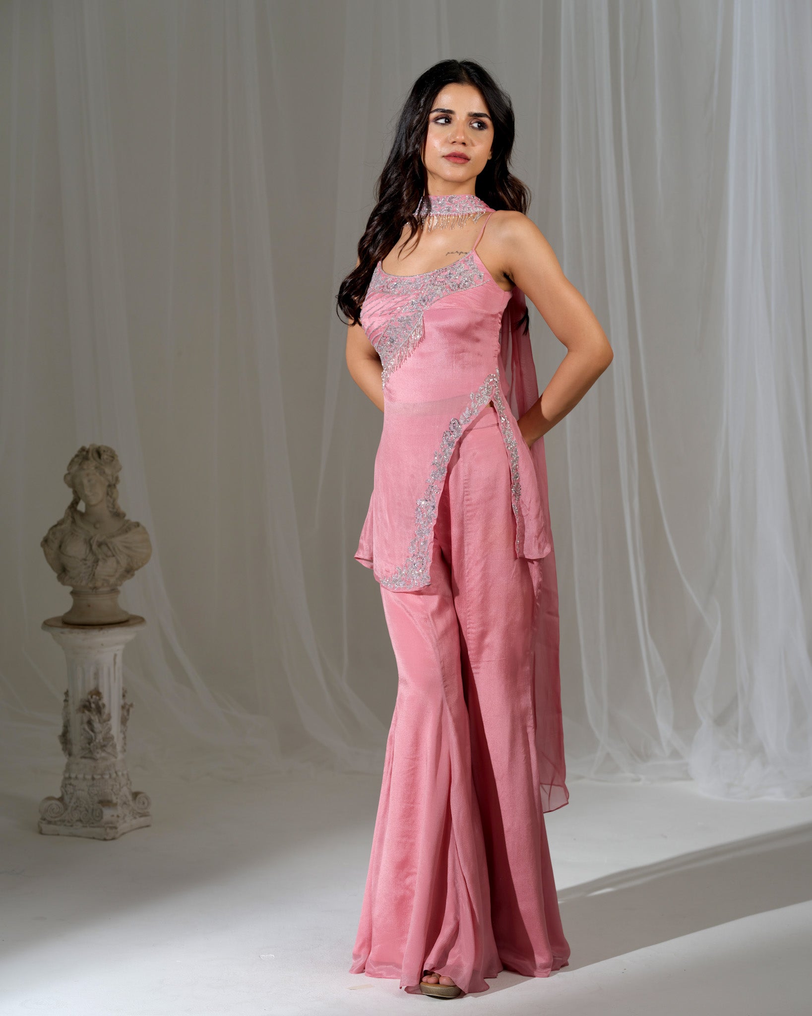 Woman wearing a pink evening gown with silver embellishments against a white curtain background.