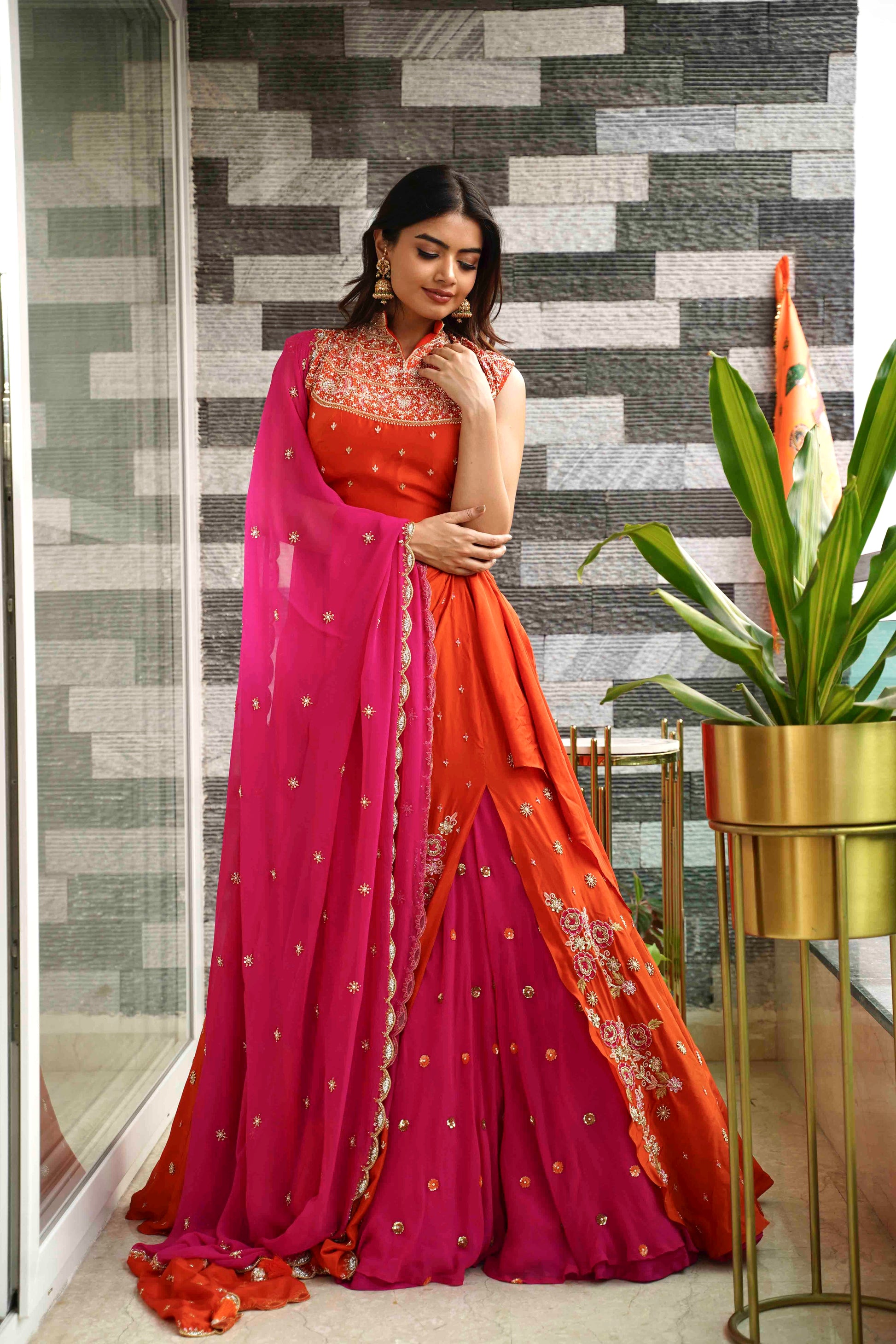 Woman in a pink and orange saree standing indoors with a patterned wall and plant in the background.