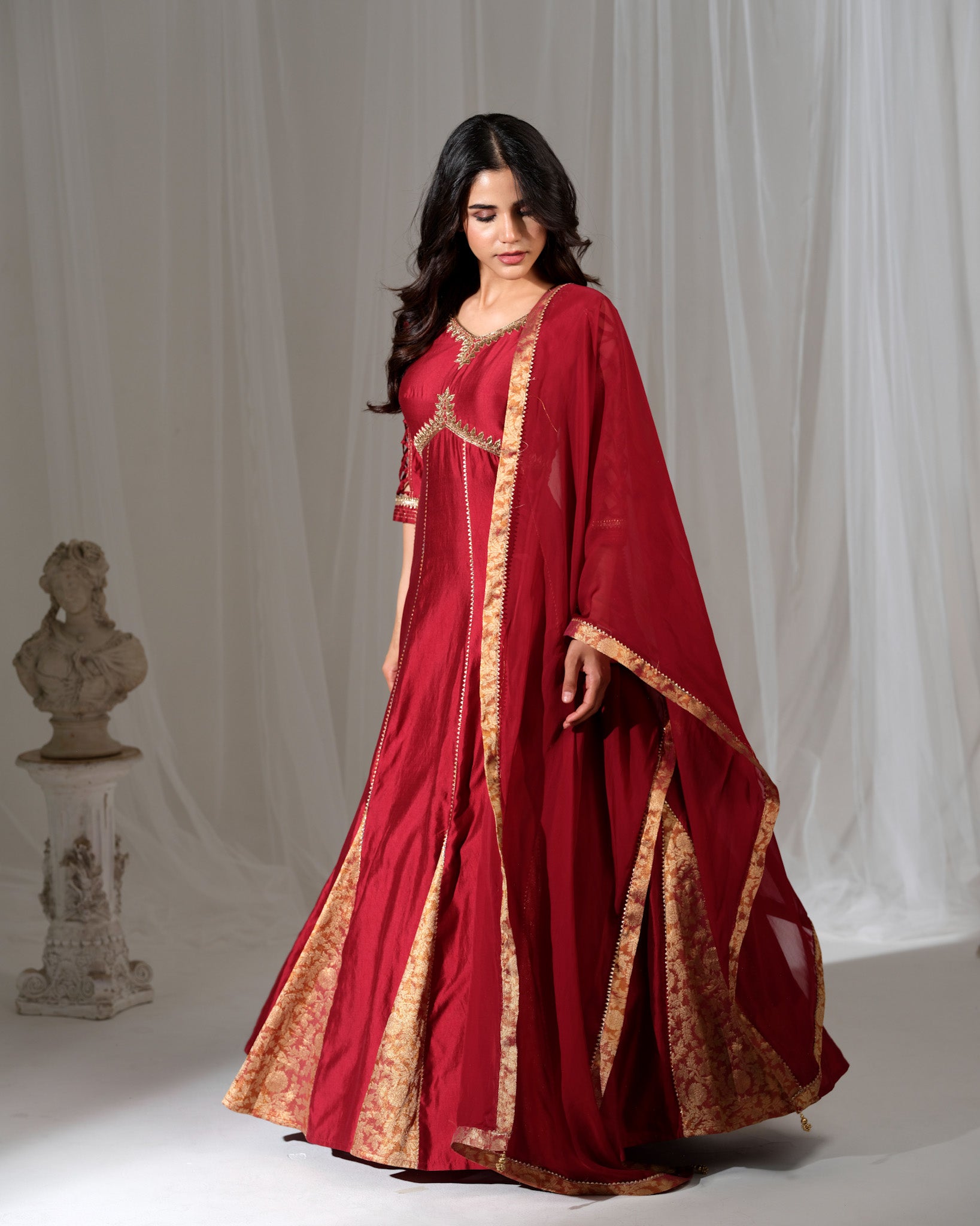 Woman wearing a red traditional outfit with gold embroidery against a white curtain background