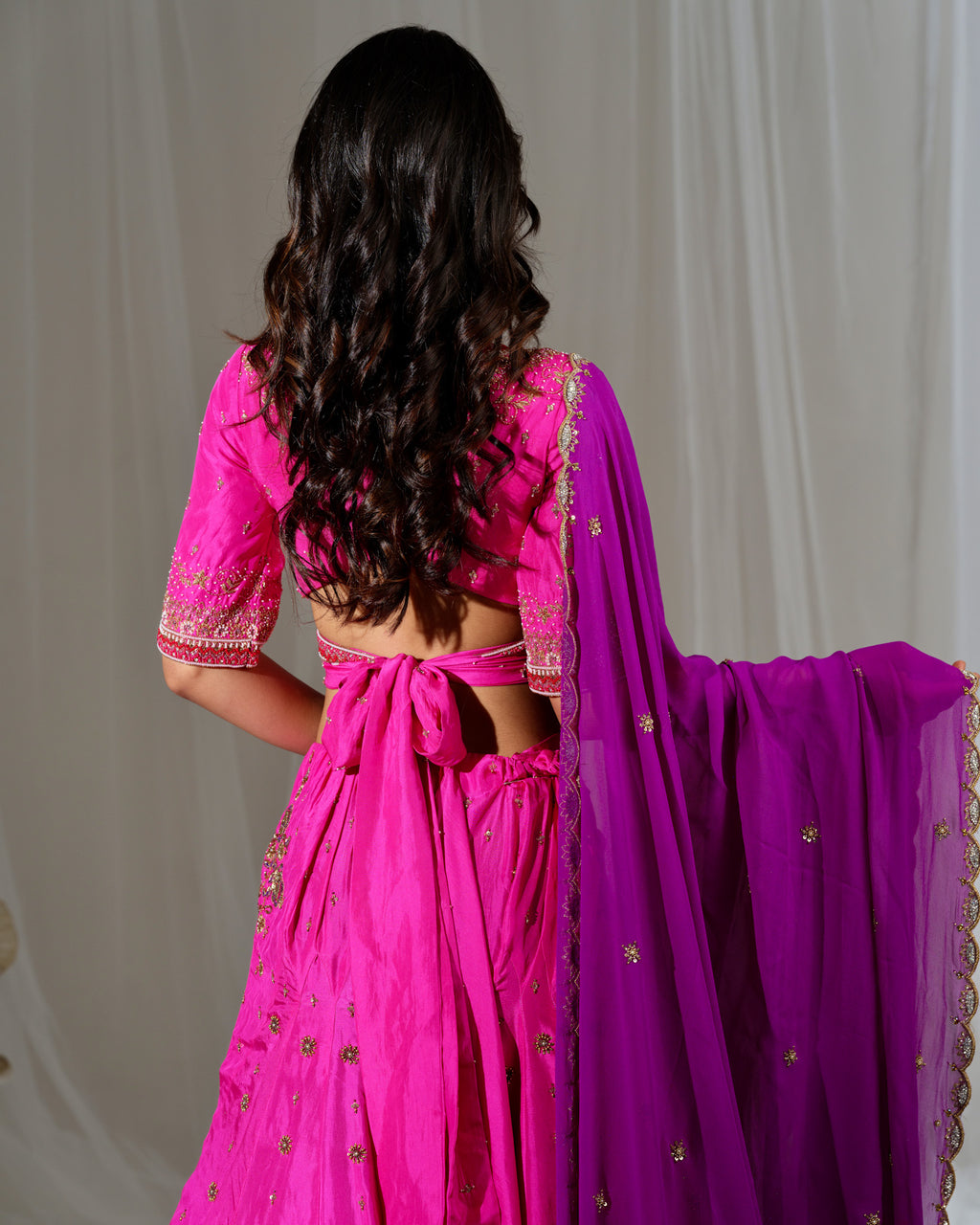 Person wearing a bright pink traditional outfit with a purple dupatta against a plain background