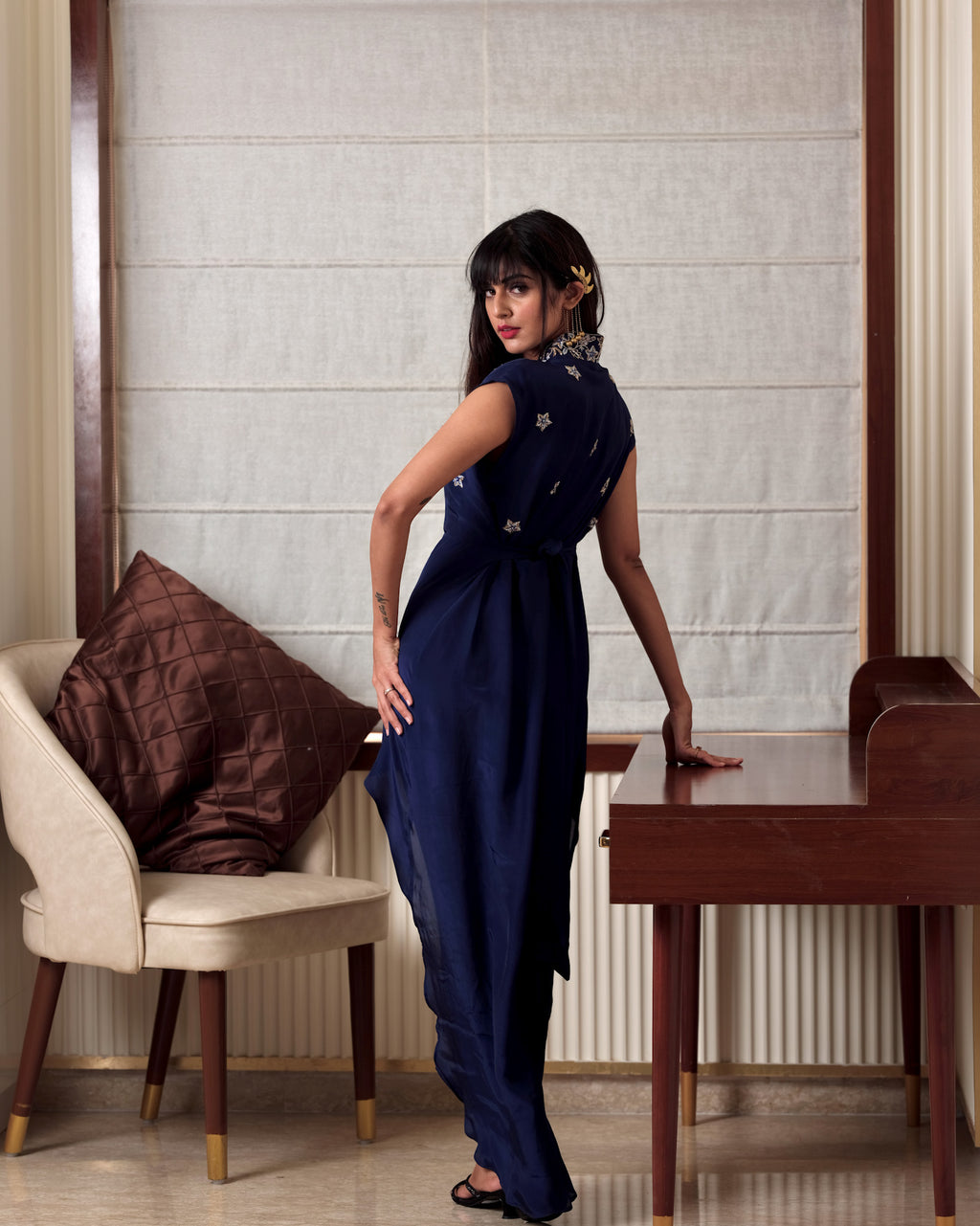 Woman in a blue dress standing in a room with a chair and table.