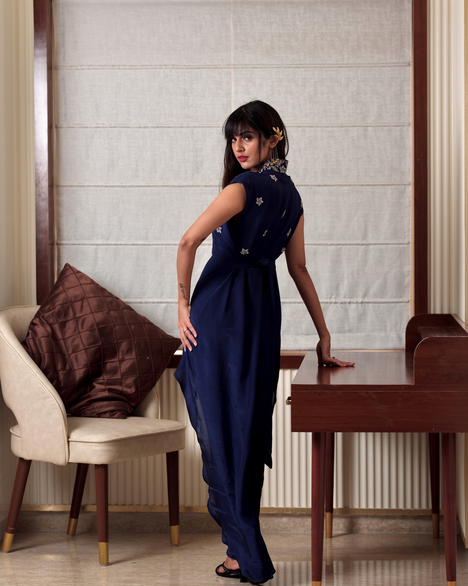 Woman in a blue dress standing in a room with a chair and table.