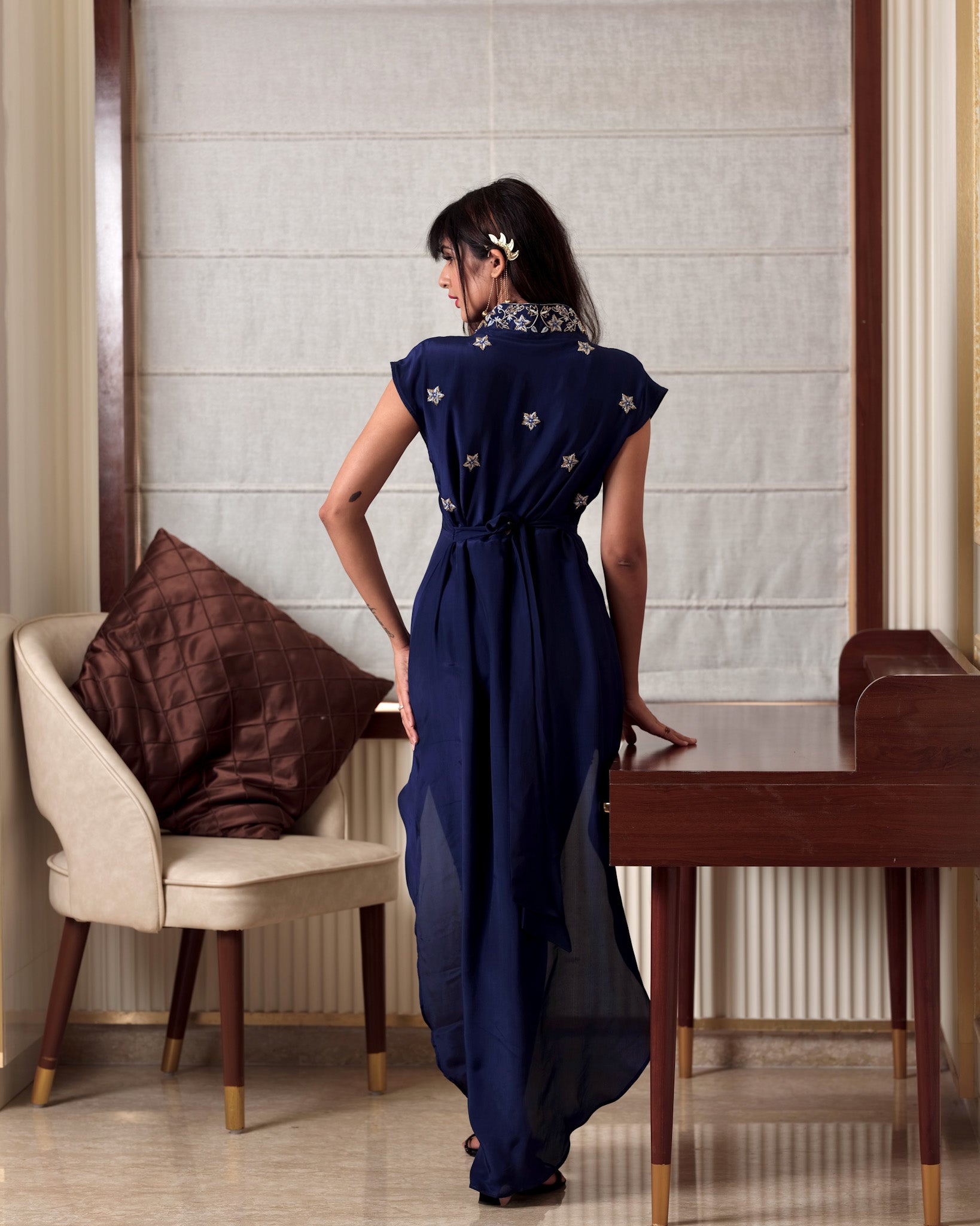 Woman in a blue dress standing in a room with a chair and table.