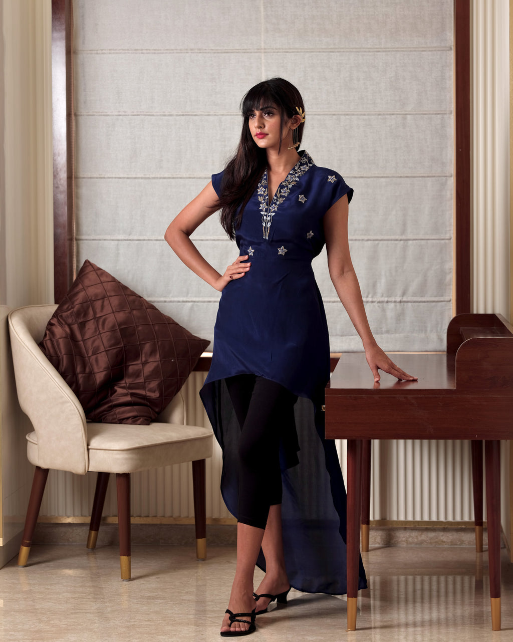 Woman in a blue dress standing in a room with a chair and table.