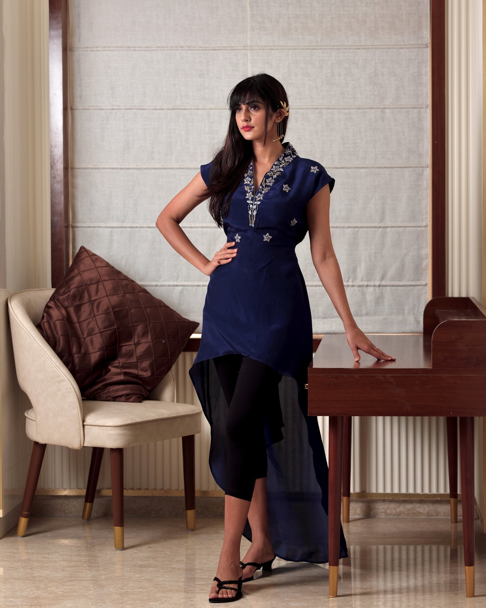 Woman in a blue dress standing in a room with a chair and table.