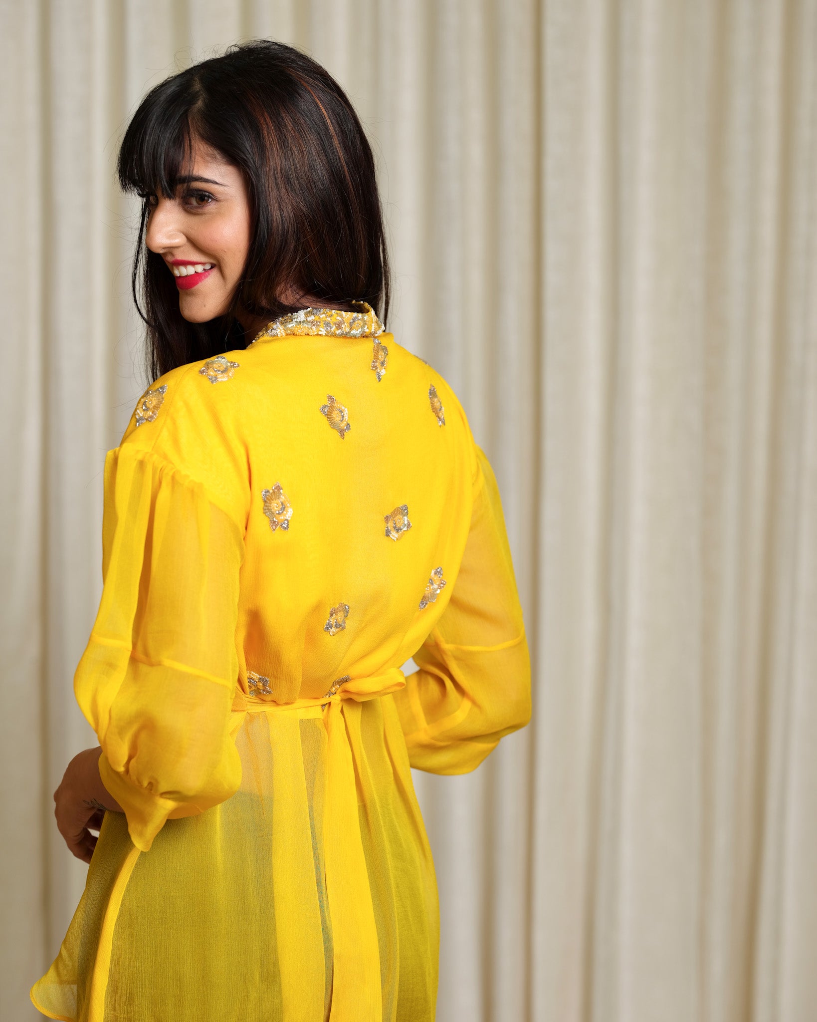 Woman wearing a yellow dress with floral patterns against a beige curtain background
