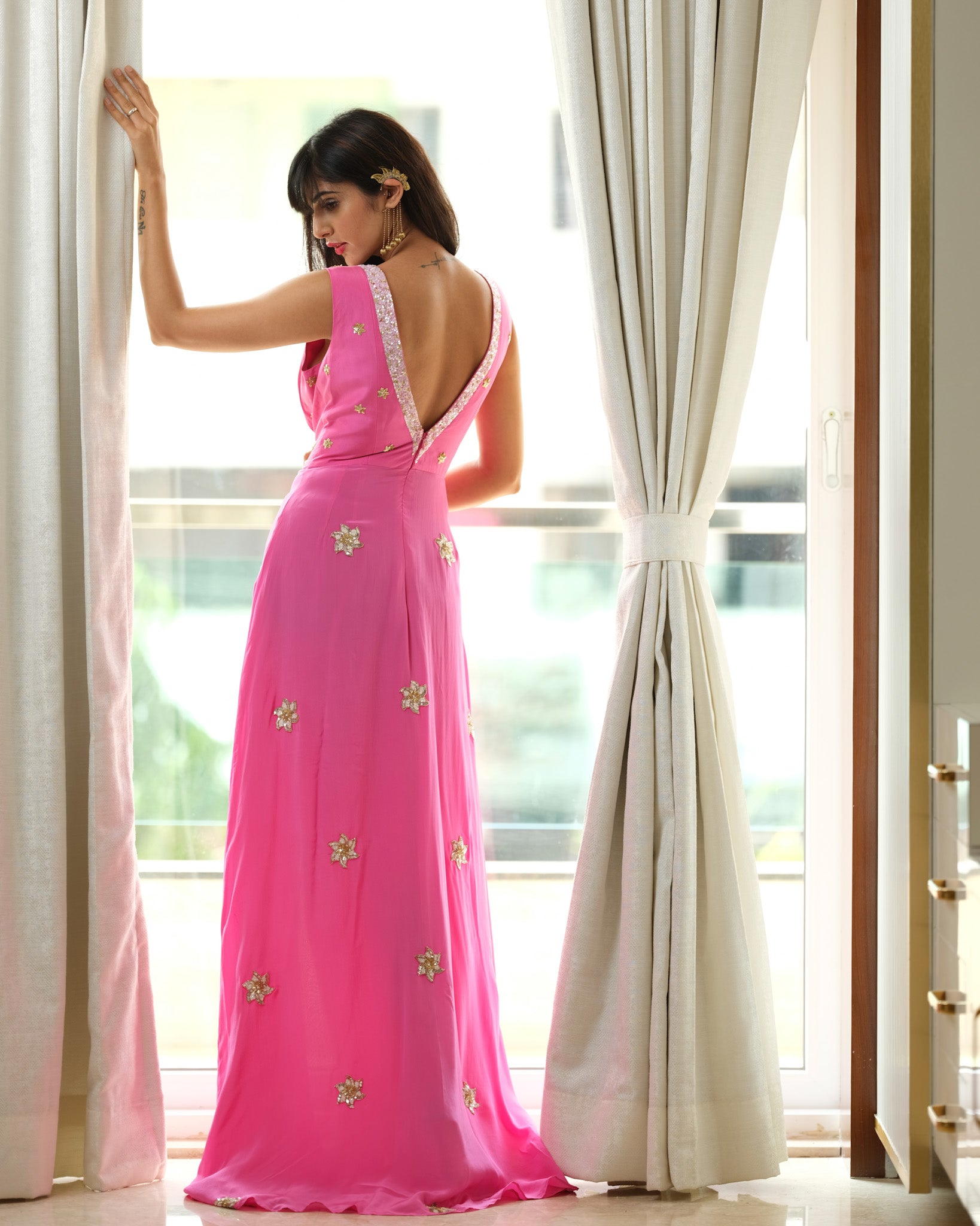 Woman in a pink evening gown with floral embellishments standing by a large window.