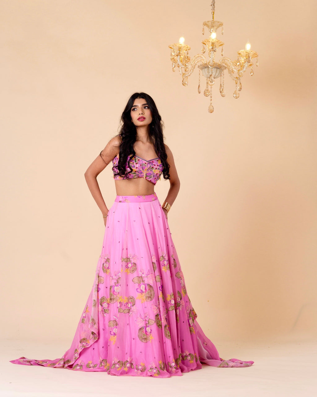 Woman wearing a pink traditional outfit with a floral top and skirt, standing against a beige background with a chandelier.