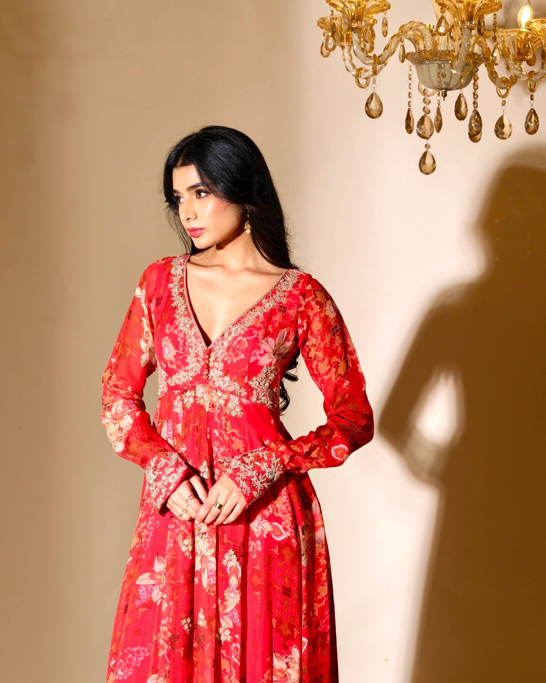 Woman in a red floral dress standing in front of a chandelier casting a shadow on the wall.