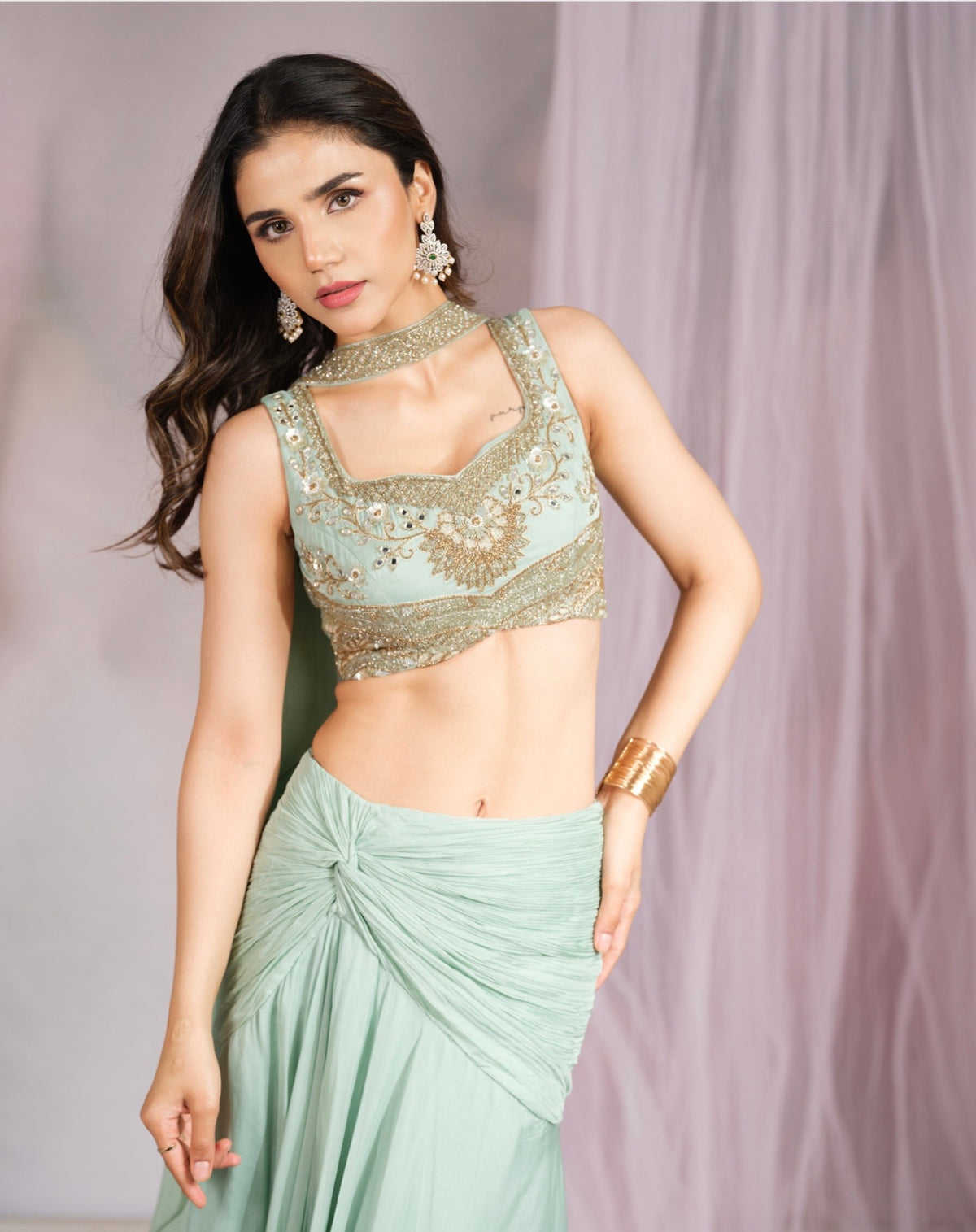 A woman modeling a mint green traditional outfit with a heavily embellished gold embroidered sleeveless blouse and a matching pleated skirt, posed against a soft lavender draped backdrop.