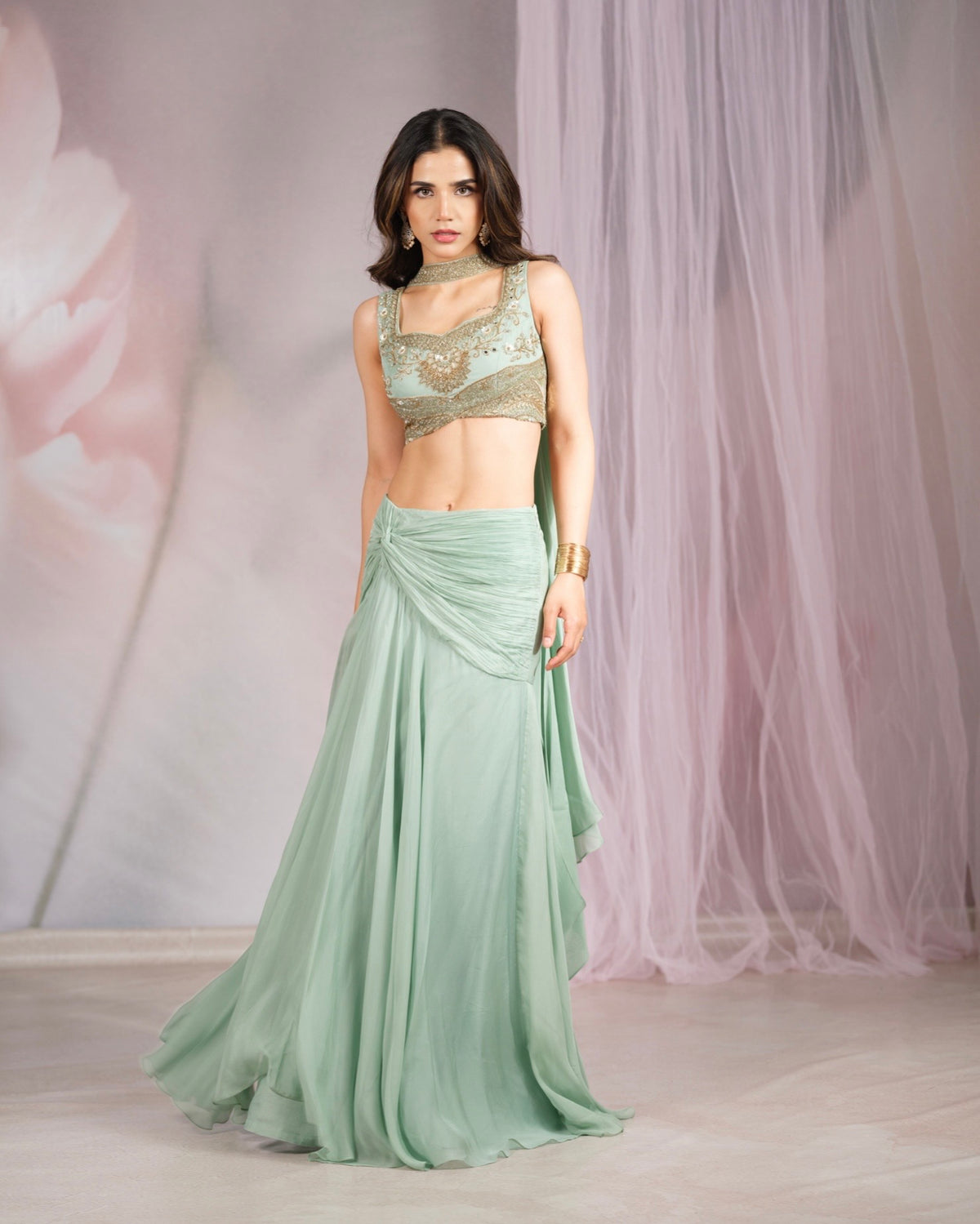 A woman modeling a mint green two-piece traditional outfit with a heavily embellished gold sequined crop top and a flowing draped skirt, set against a soft pink tulle backdrop.