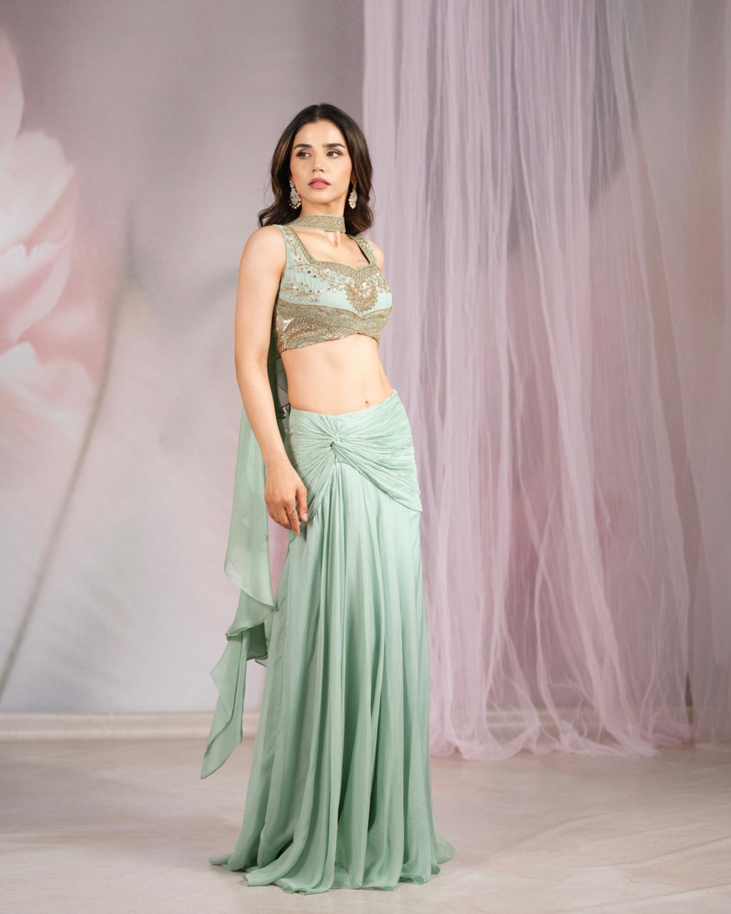 A woman models a mint green two-piece traditional outfit featuring a sleeveless draped skirt and a matching sheer draped scarf, paired with a gold embroidered crop top. She stands in front of a soft pink tulle curtain backdrop.