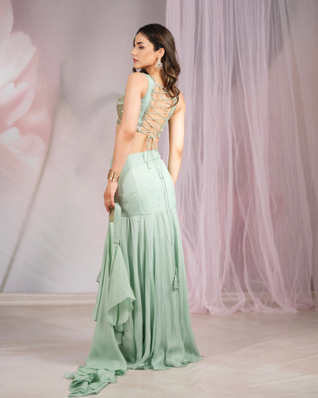 A woman modeling a mint green maxi dress with a lace-up open back, shown from the back in a studio setting with pink tulle drapes.