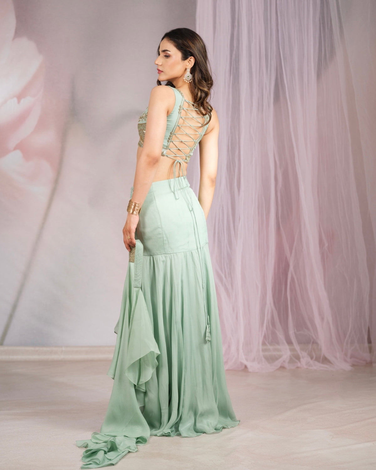 A woman modeling a mint green maxi dress with a lace-up open back, shown from the back in a studio setting with pink tulle drapes.