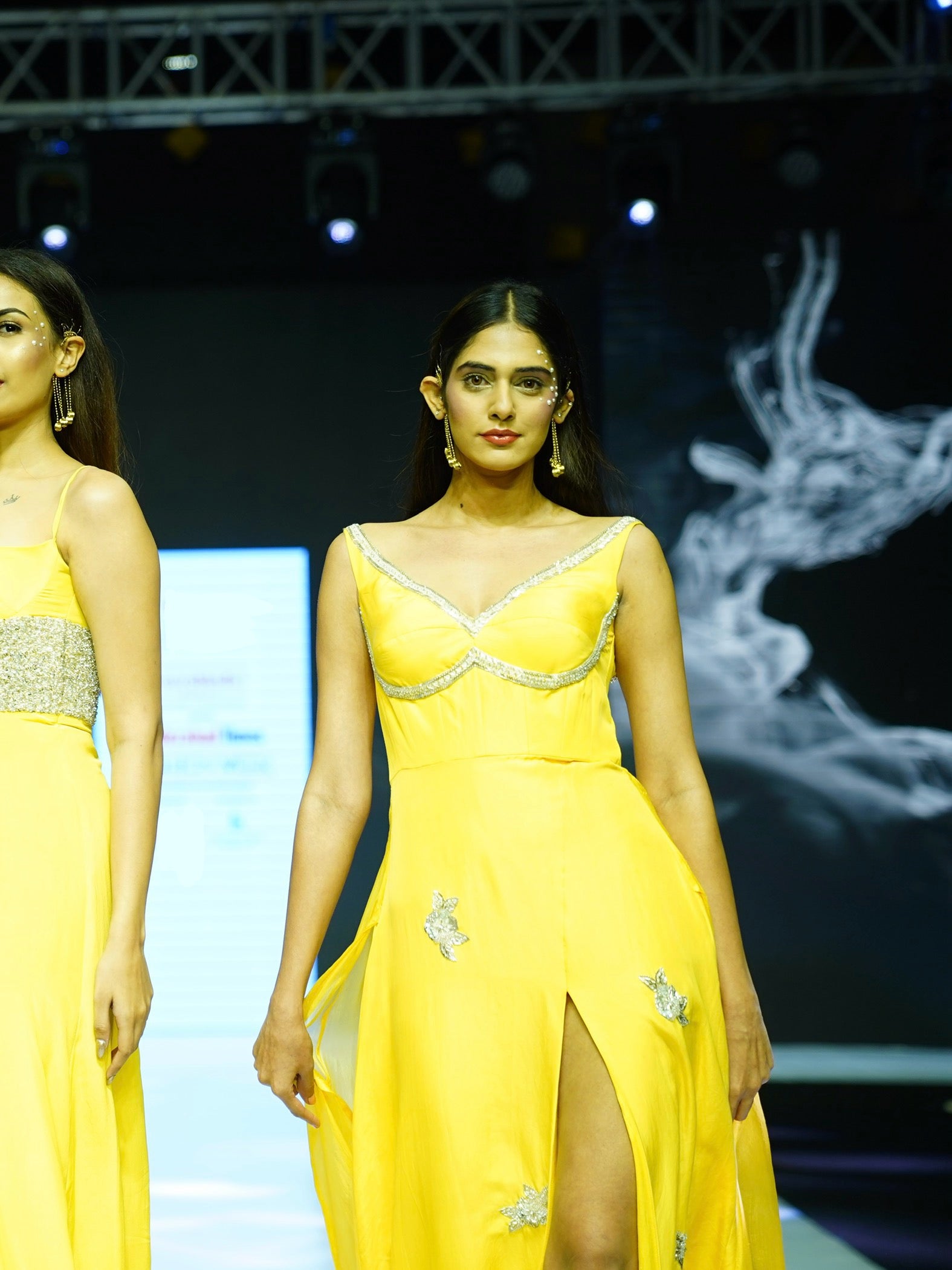 Model wearing a yellow evening gown with intricate detailing on a fashion show runway.