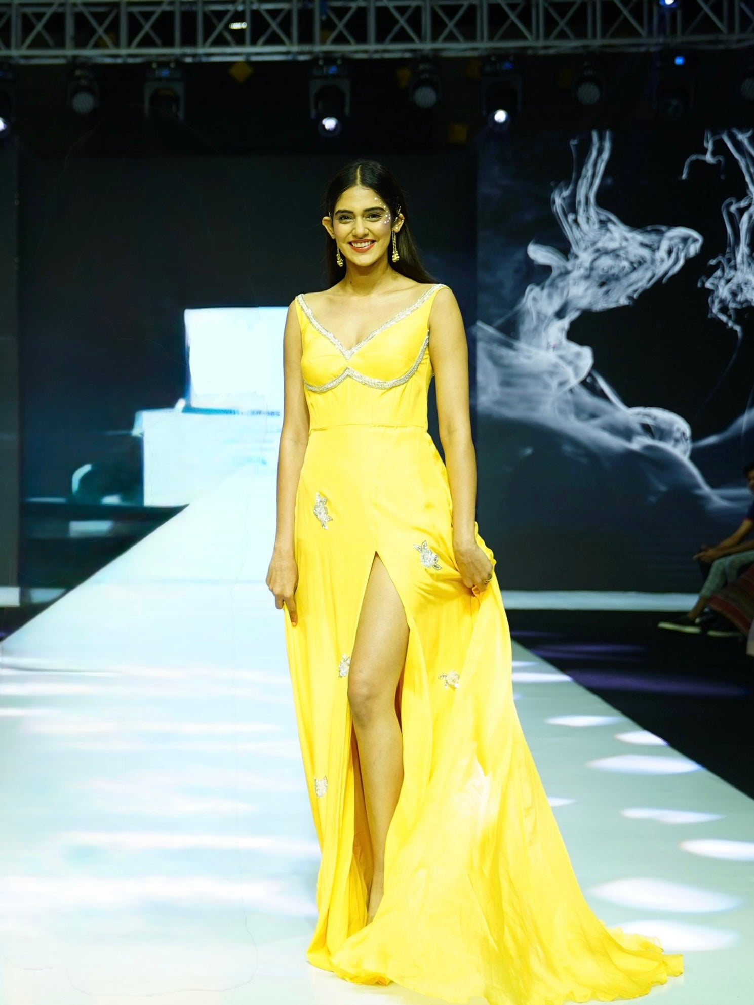 Model wearing a yellow evening gown on a runway with a dark background