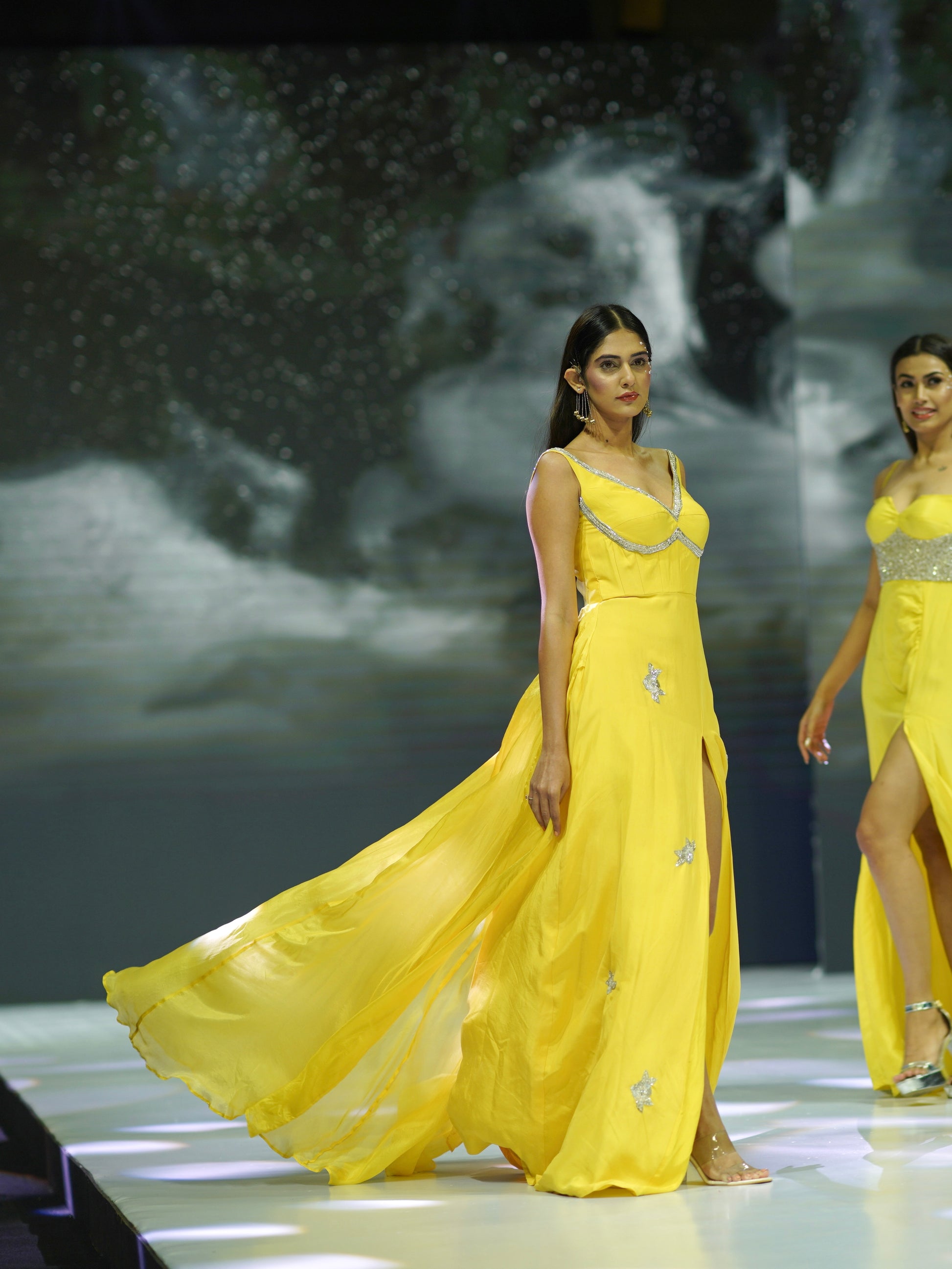 Two models in yellow evening gowns on a runway with a dark, abstract background.