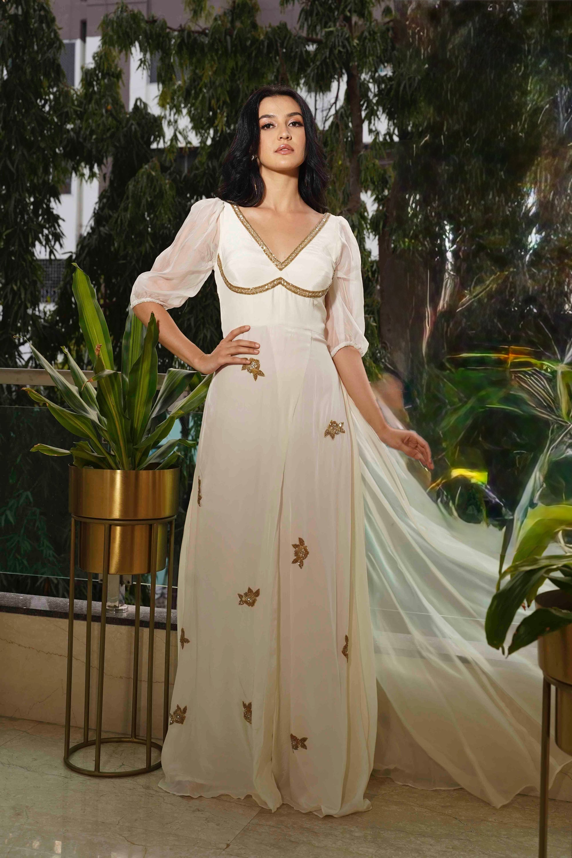 Woman in a white gown with gold embroidery standing among plants.