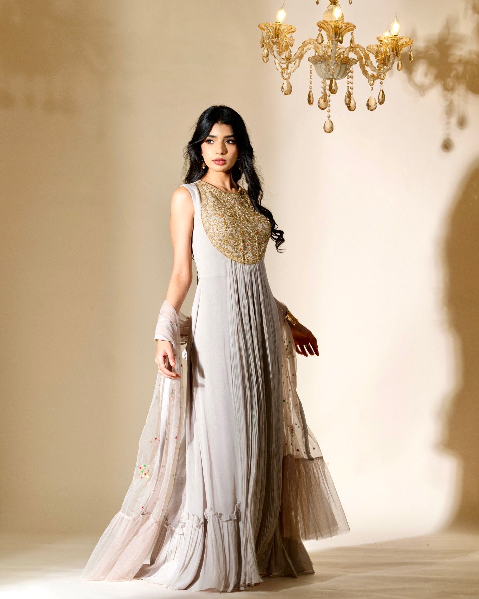 Woman in a white and gold traditional outfit standing in a softly lit room with a chandelier.