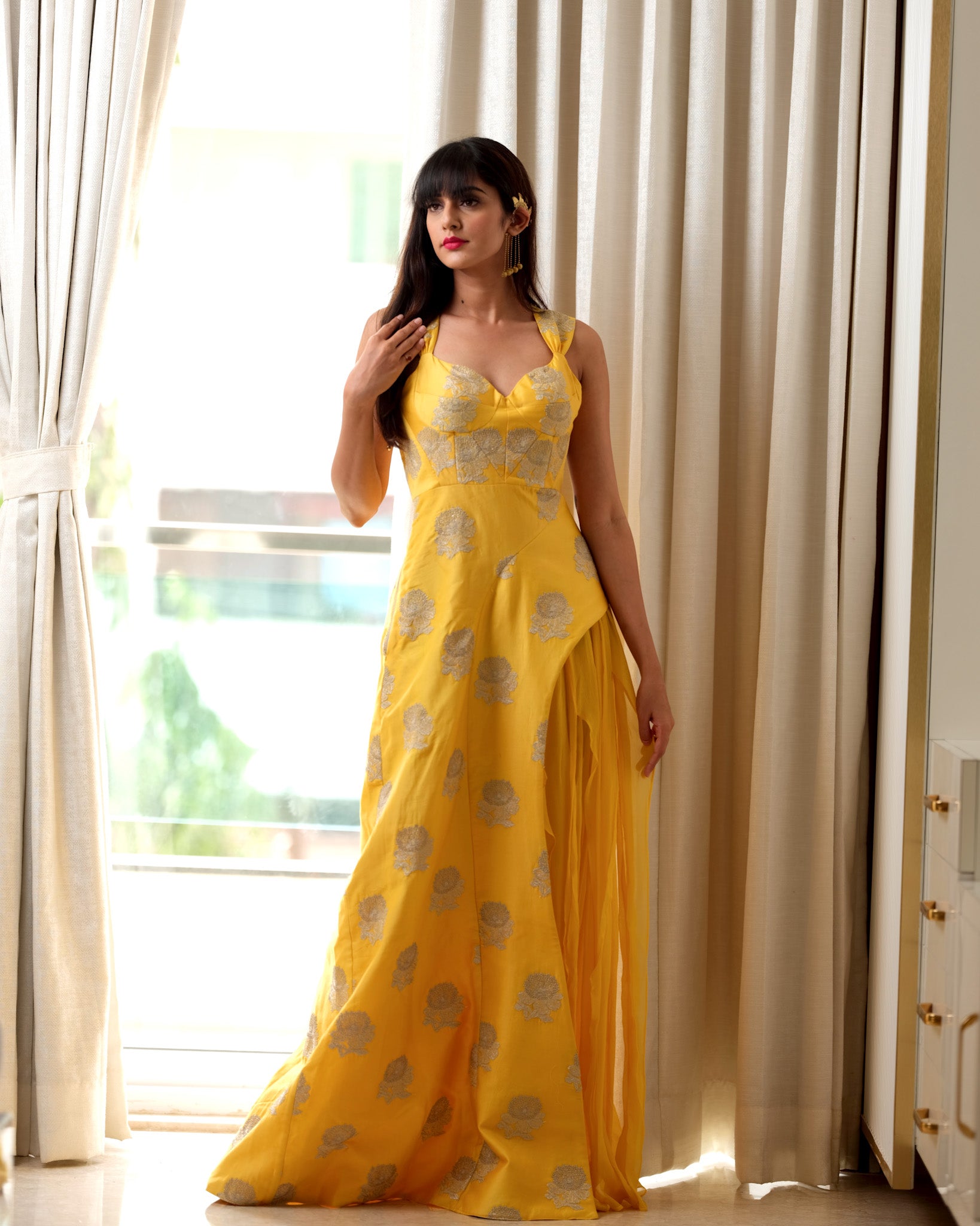 Woman wearing a yellow floral dress standing in a bright room with white curtains.