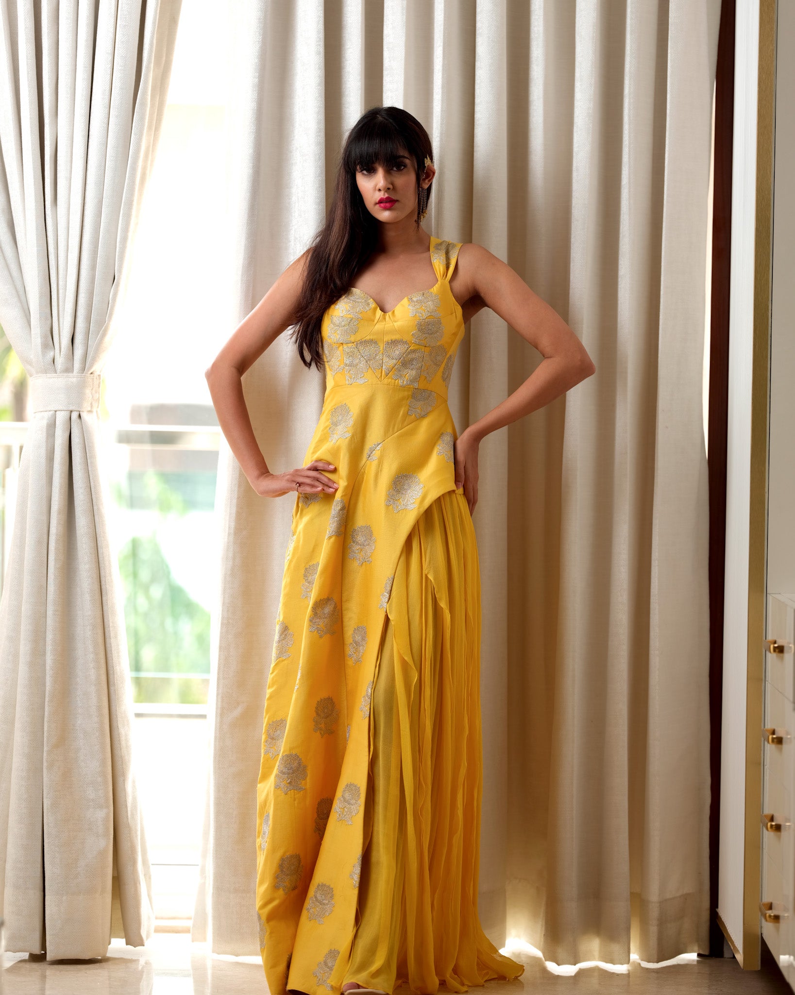 Woman wearing a yellow dress with patterns standing in front of white curtains.