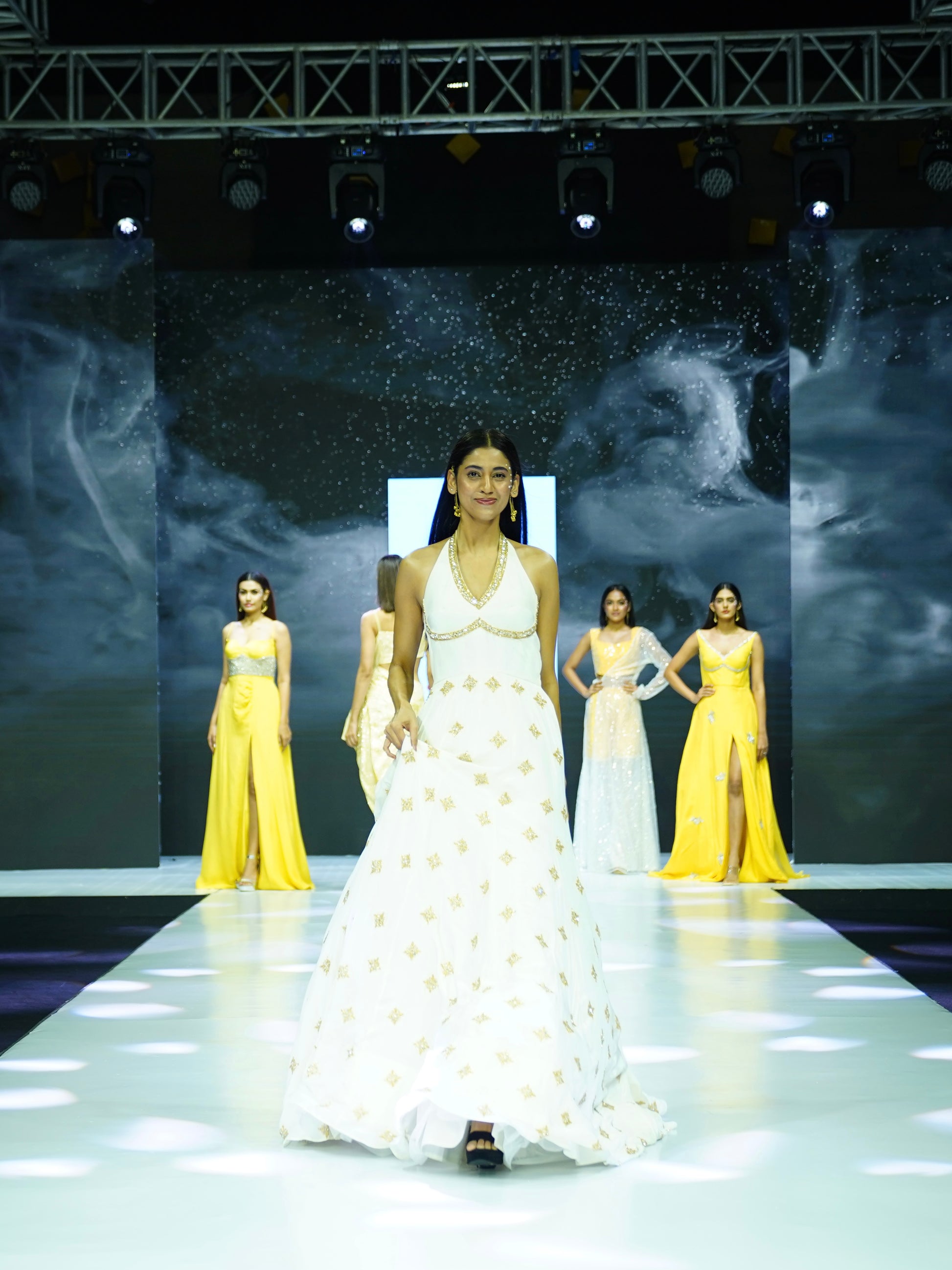 Model walking on a fashion show runway wearing a white dress with yellow accents.