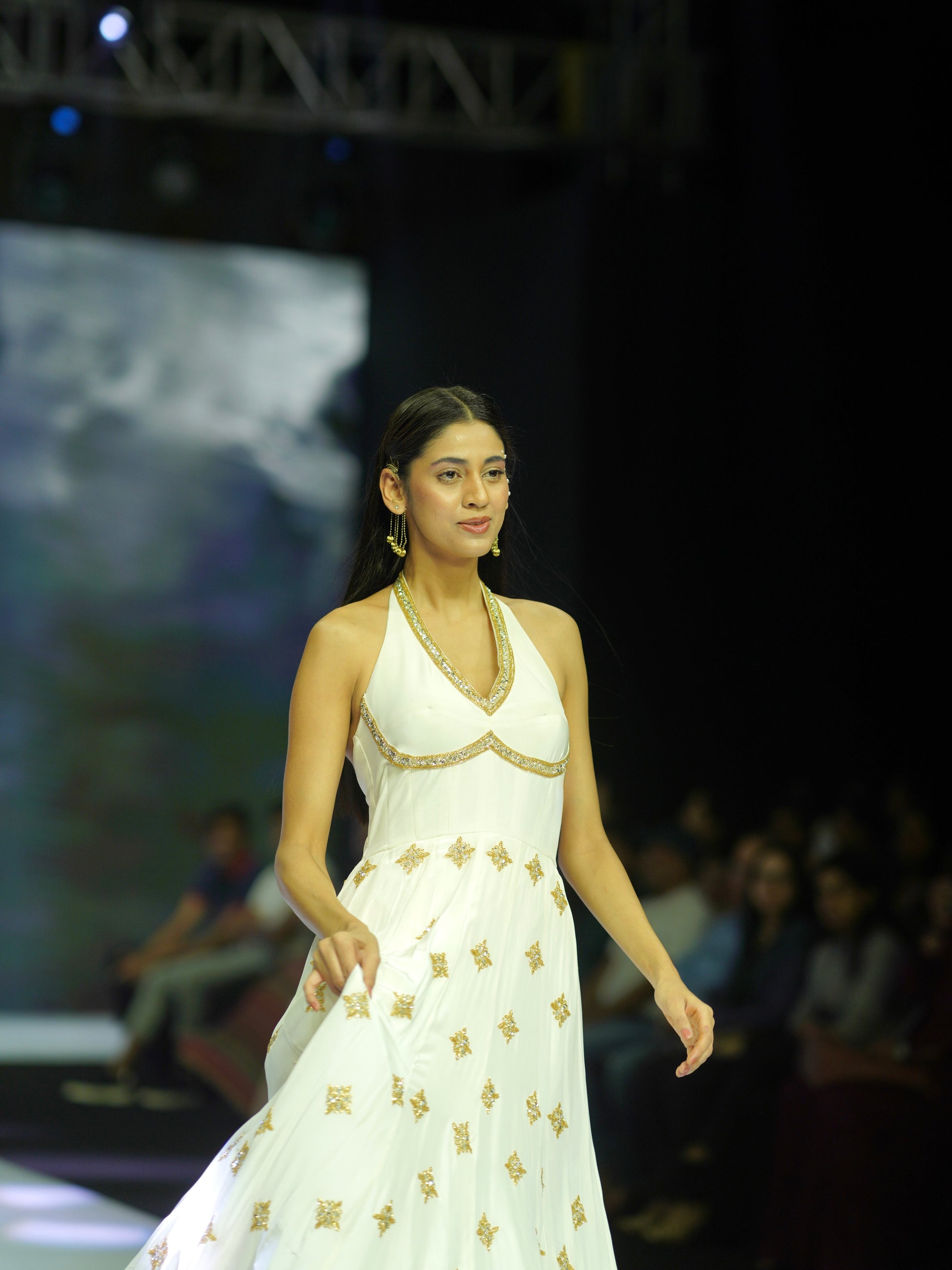 Model wearing a white dress with gold patterns on a fashion show runway.