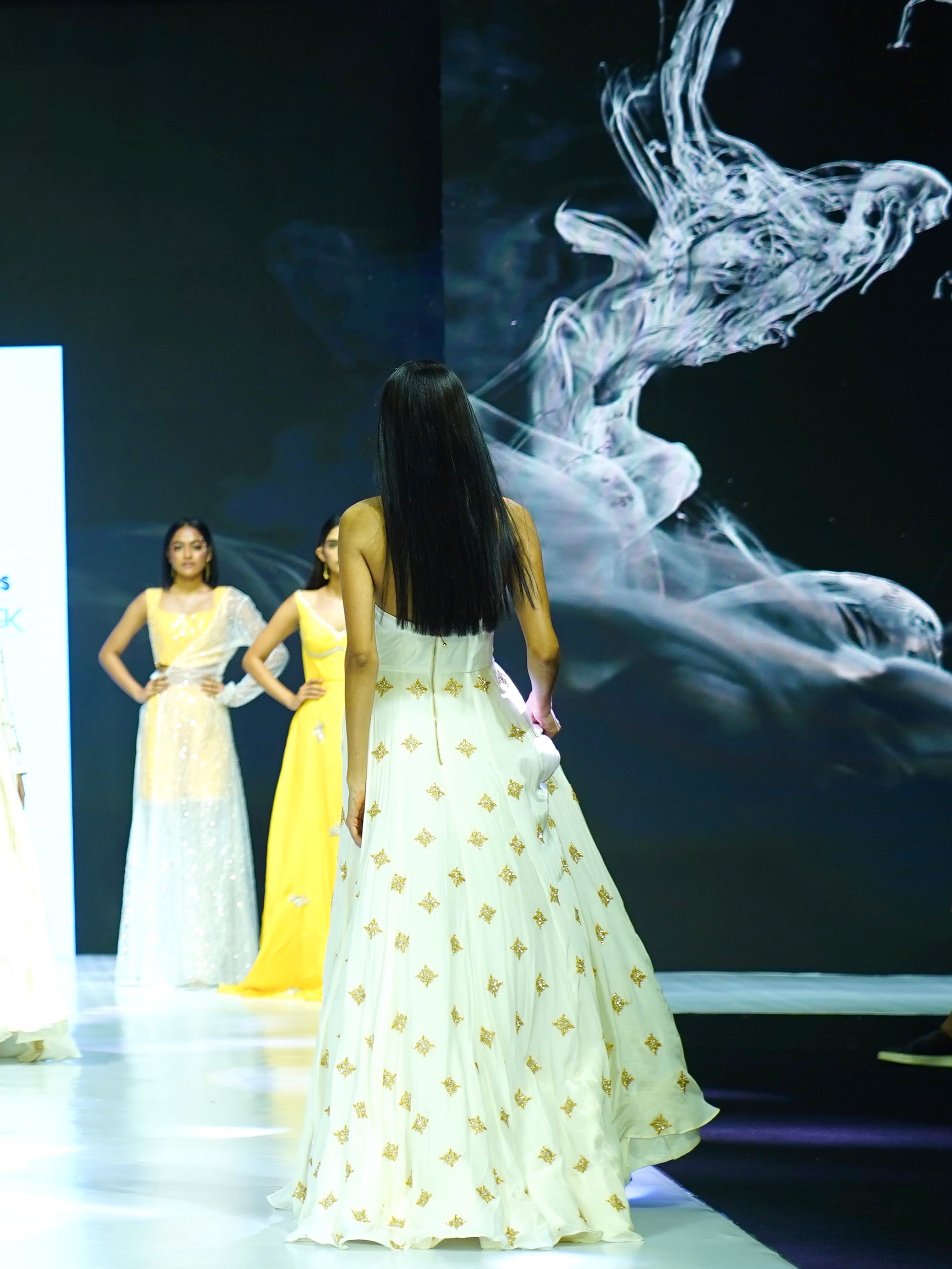 Two models on a runway wearing white dresses with gold patterns, with a large screen displaying abstract smoke art in the background.